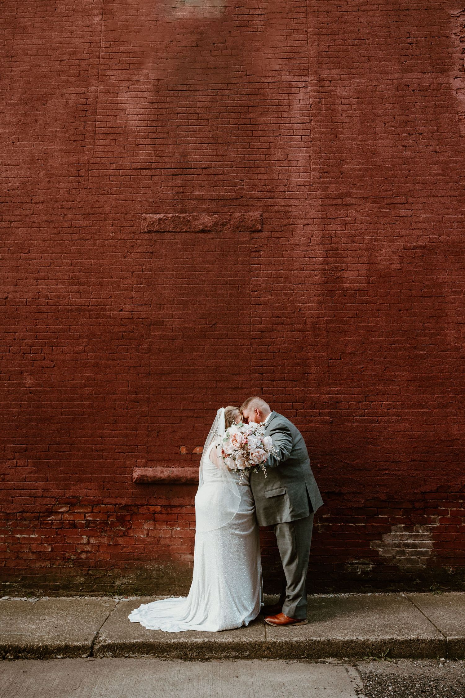 Wedding couple embracing against weathered red brick wall.