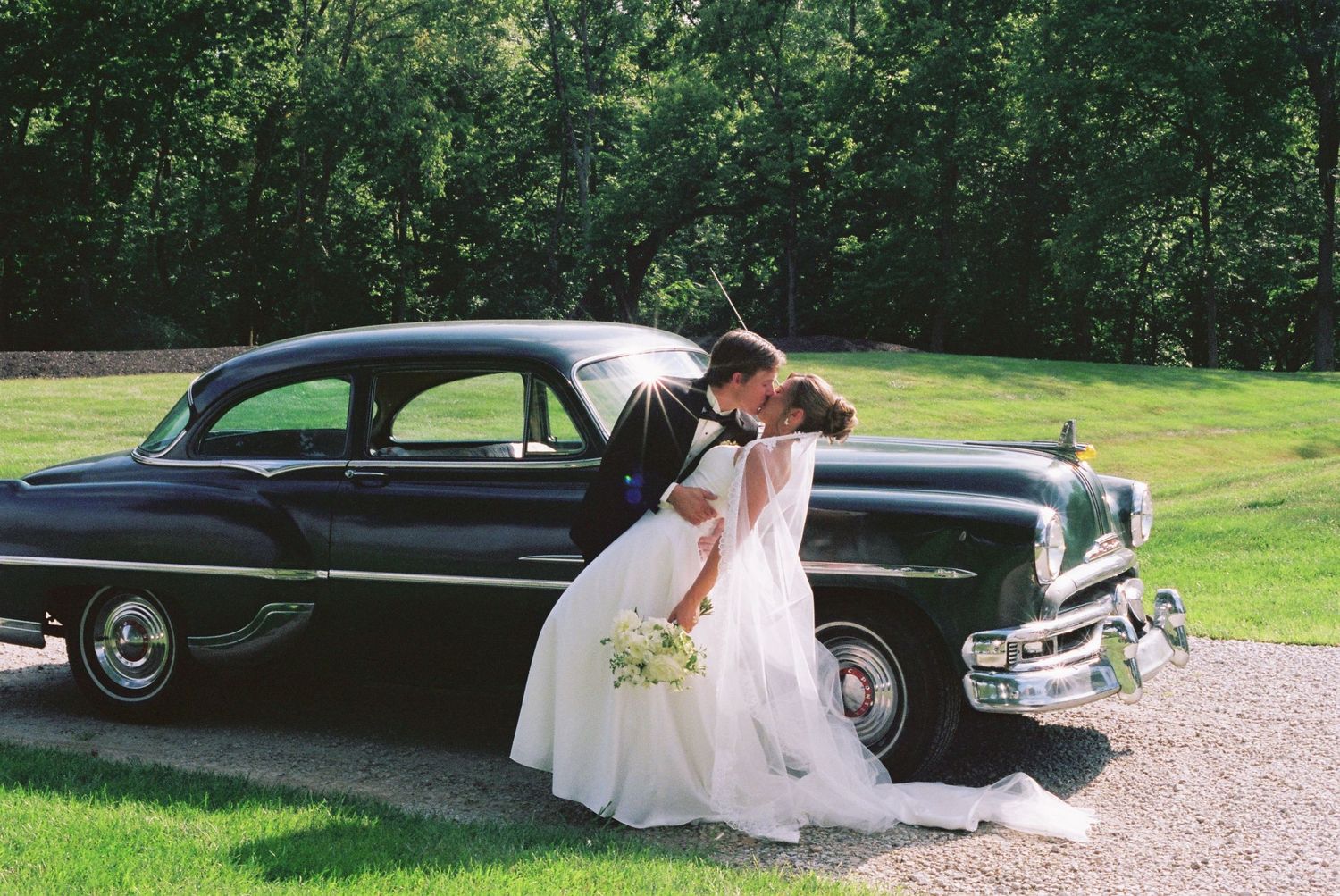 Bride and groom sharing a kiss beside a classic black vintage car on their wedding day.