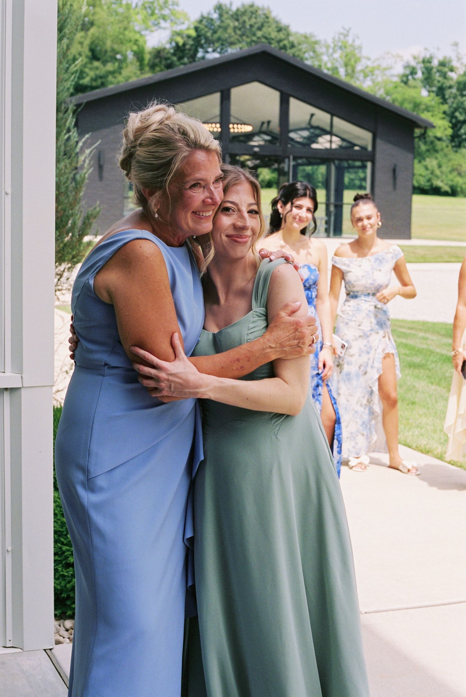 Two women in elegant gowns share a warm hug at an outdoor wedding venue with guests approaching in background.