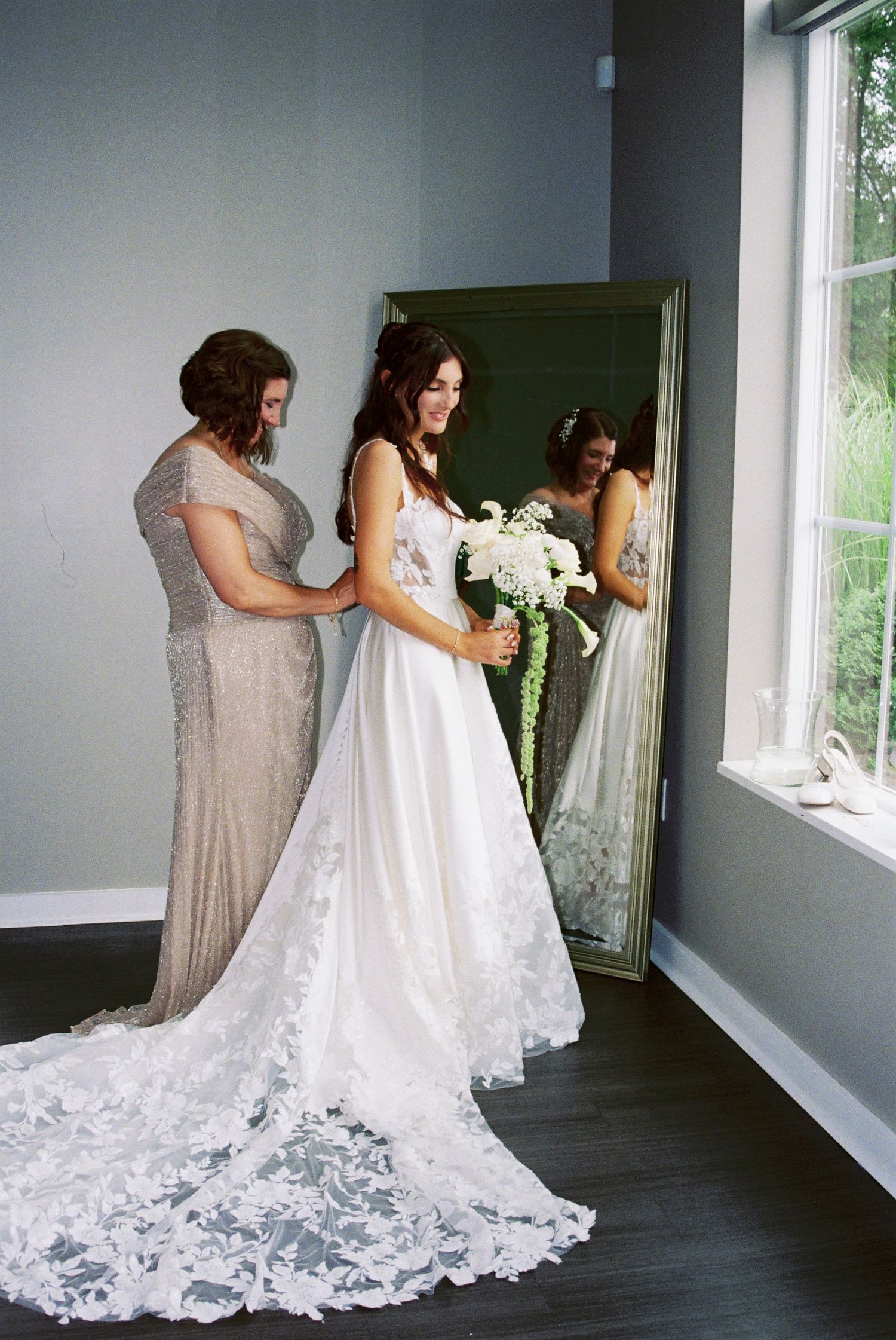 Bride in elegant lace wedding gown holds white bouquet while bridesmaid fastens dress by mirror.