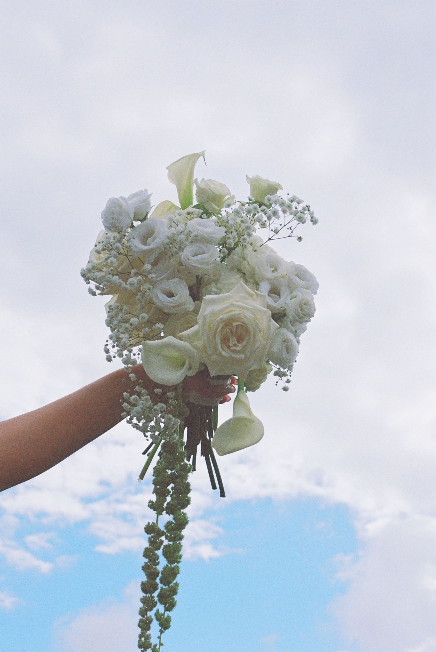 Hand holding a lush white bridal bouquet of roses, calla lilies, and baby's breath against a cloudy sky.