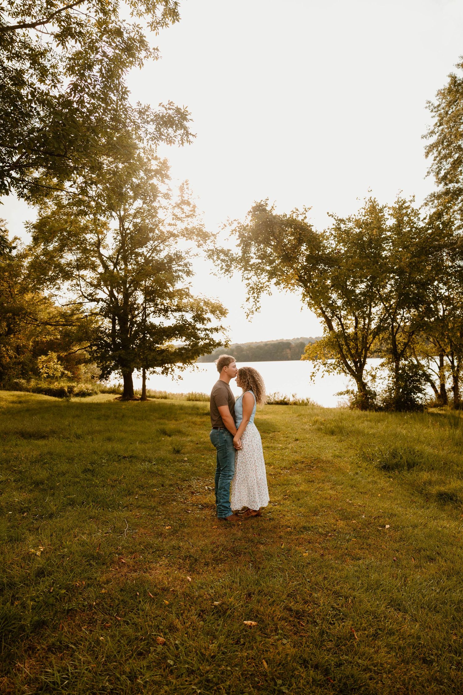 Couple standing together on grassy hill with trees and water in background during sunset.