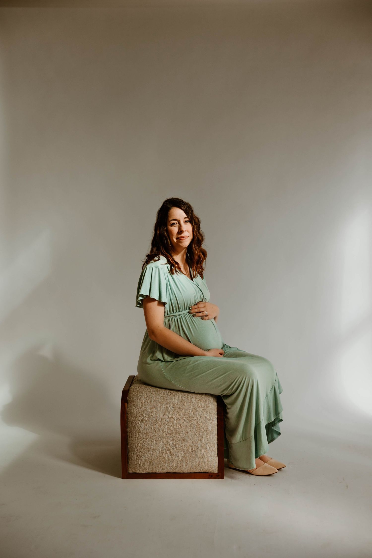 A maternity photo shows an expectant mother in a sage green dress posing on a brown chair.