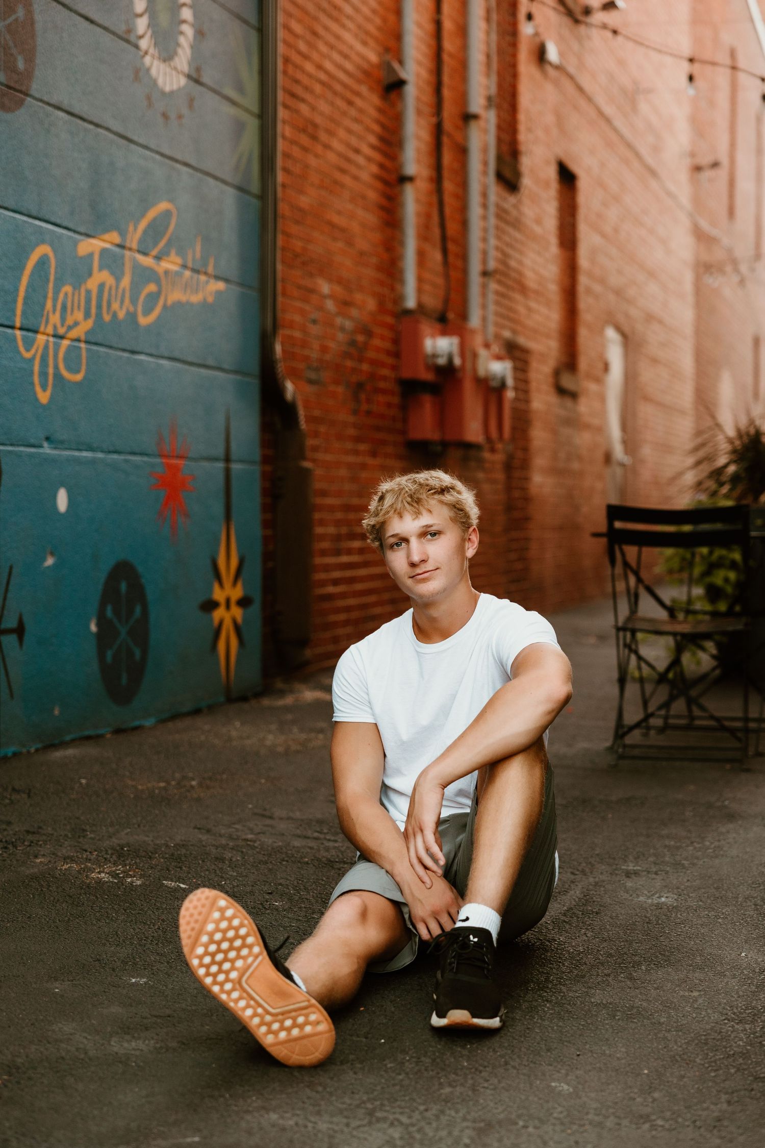 Casual street style photo showing relaxed seated pose against colorful alley wall.