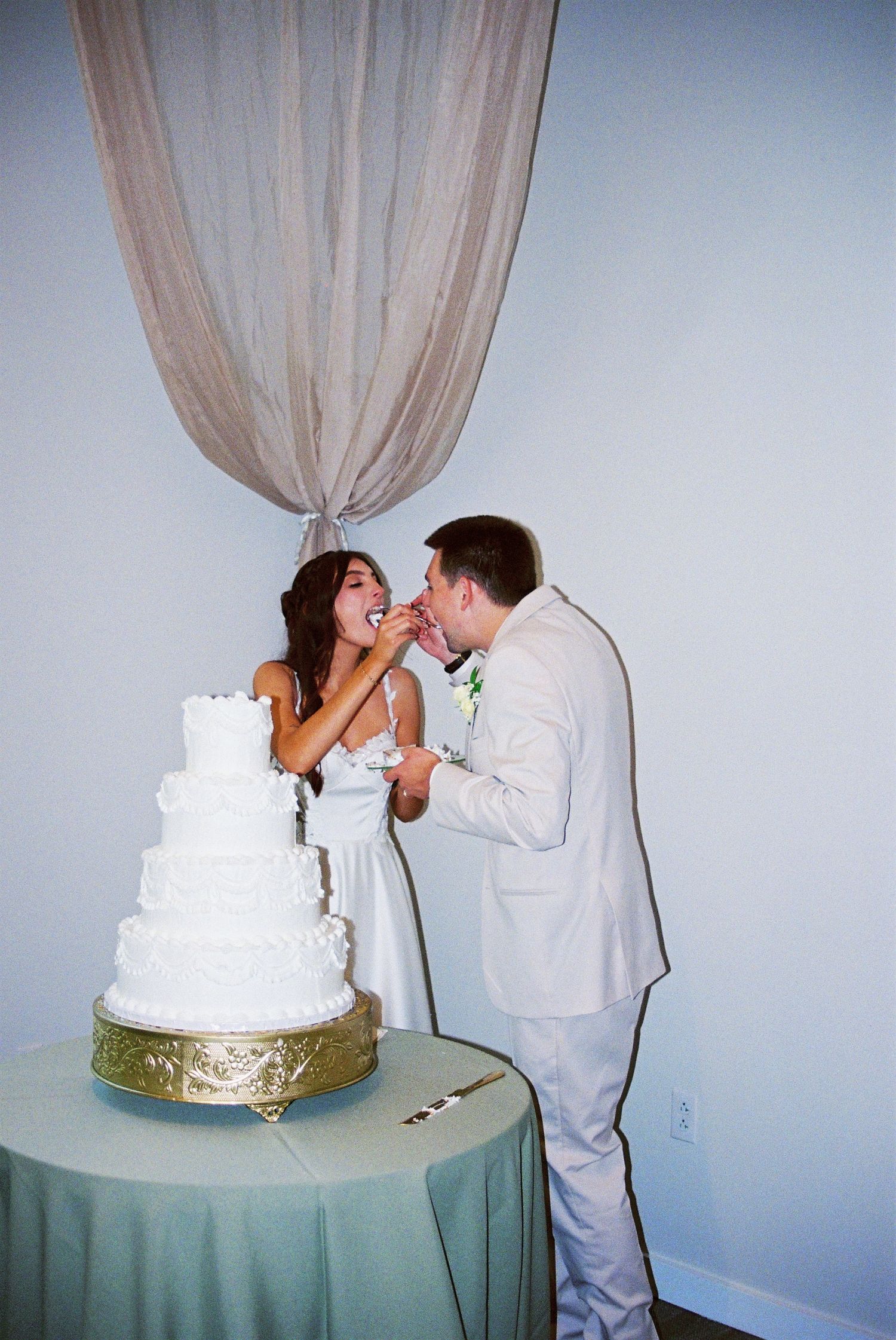 Bride and groom share playful cake tasting moment beside white four-tier wedding cake at reception.