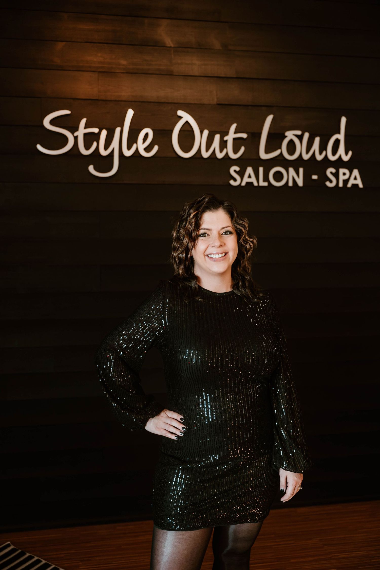 Glamorous portrait in sparkly black dress against wooden wall with salon signage.
