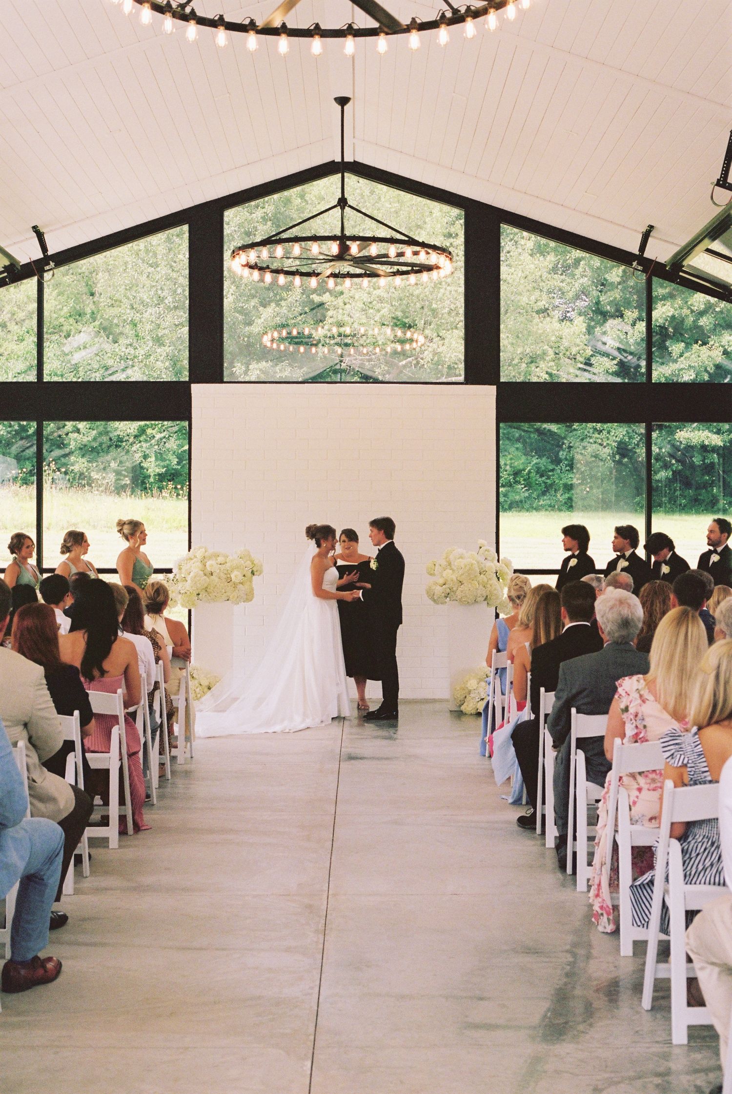 Elegant indoor wedding ceremony in a modern barn venue with white chairs, floral arrangements, and large windows overlooking greenery.