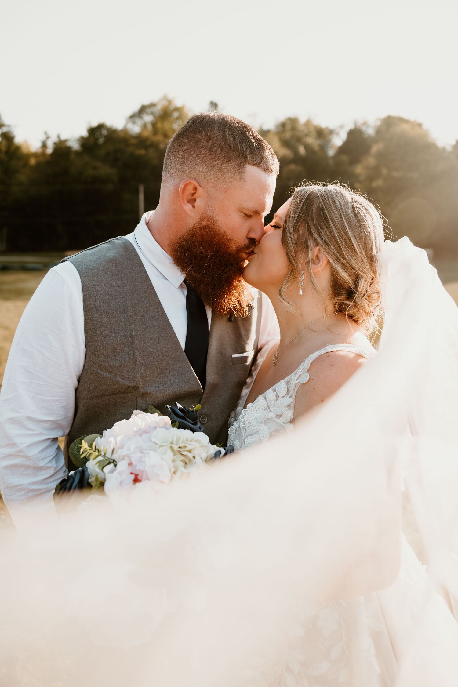 Romantic wedding couple shares intimate moment during golden hour with flowing veil in foreground.