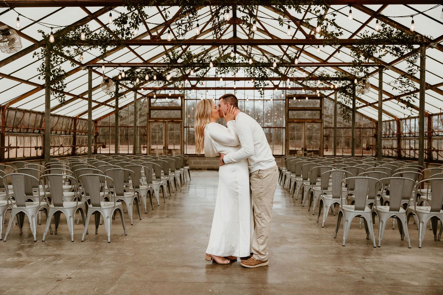 A couple shares a tender moment in an industrial greenhouse venue with metal chairs lined up for an event.