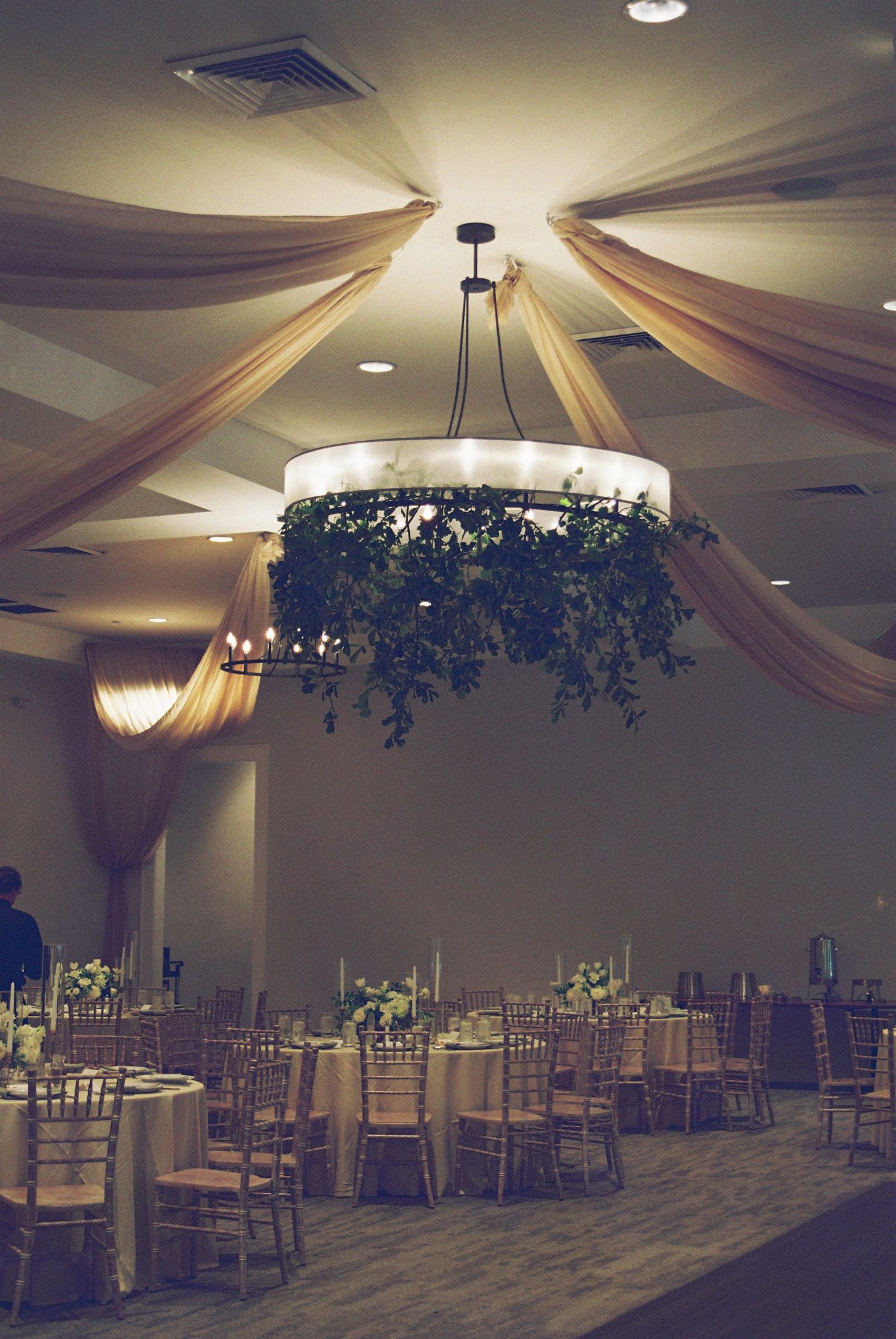 Elegant wedding reception hall with draped ceiling, greenery chandelier, and gold chiavari chairs at round tables.