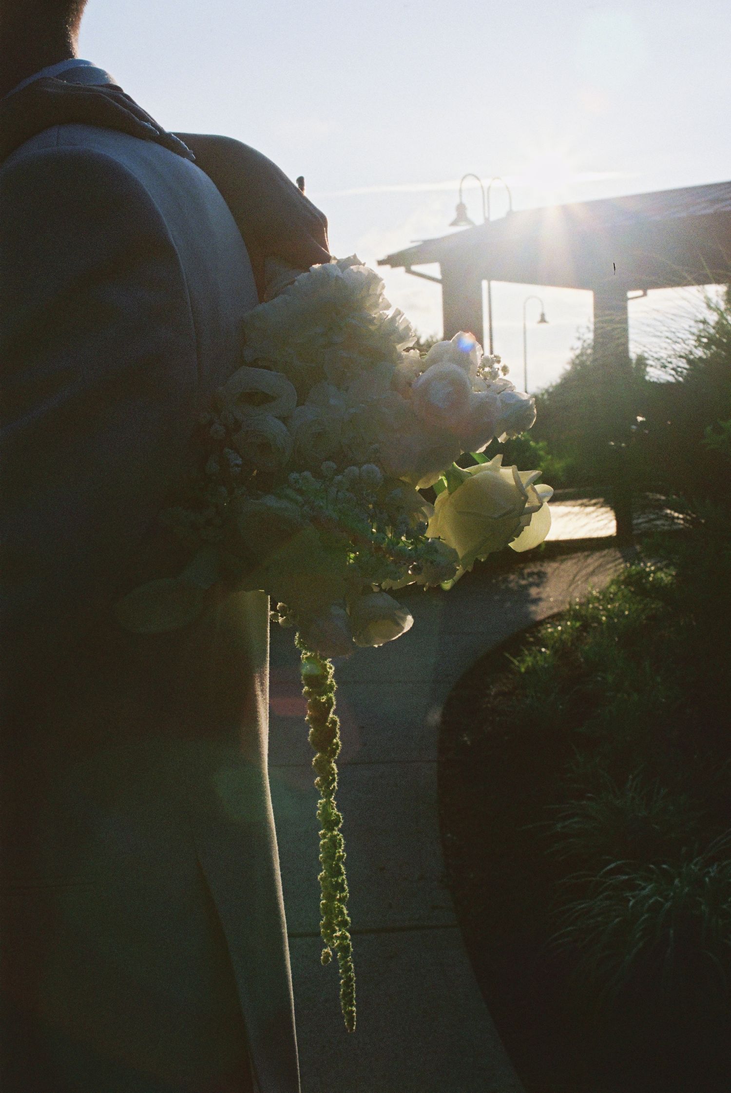 Silhouette of bride holding white bouquet backlit by golden sunlight at outdoor wedding venue.
