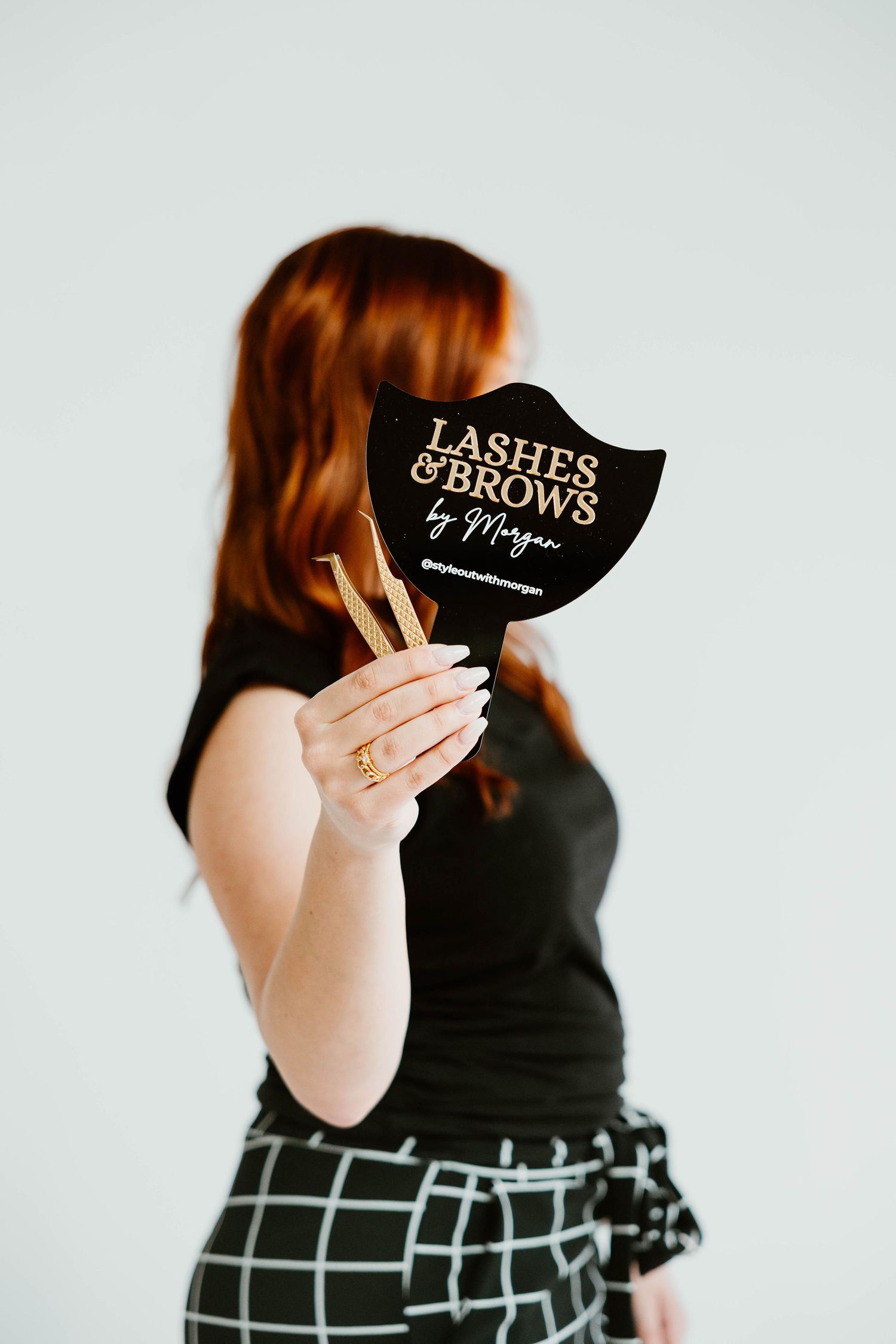 Person holding black 'Lashes & Brows' sign wearing checkered skirt in studio.
