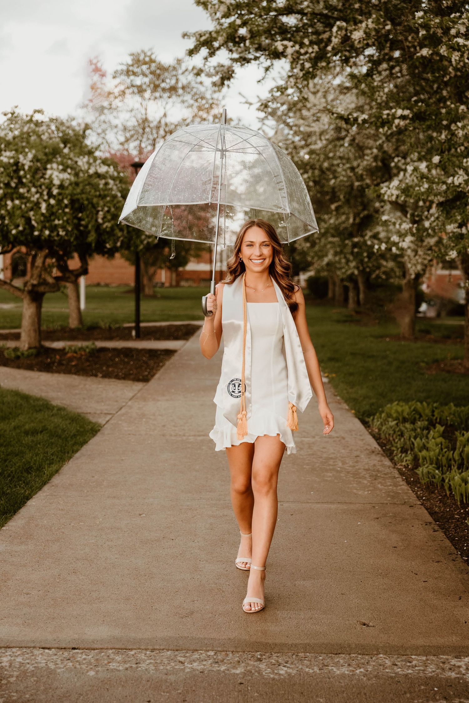 A person in a white dress walks down a sidewalk holding a clear umbrella on a sunny day.