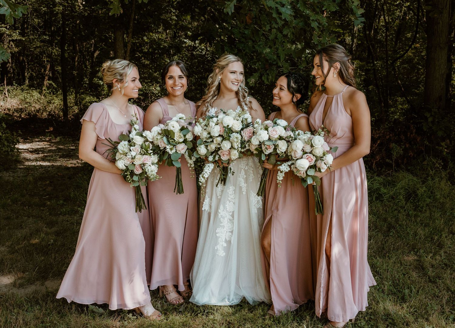 Bridesmaids in pink dresses holding white floral bouquets stand together outdoors.