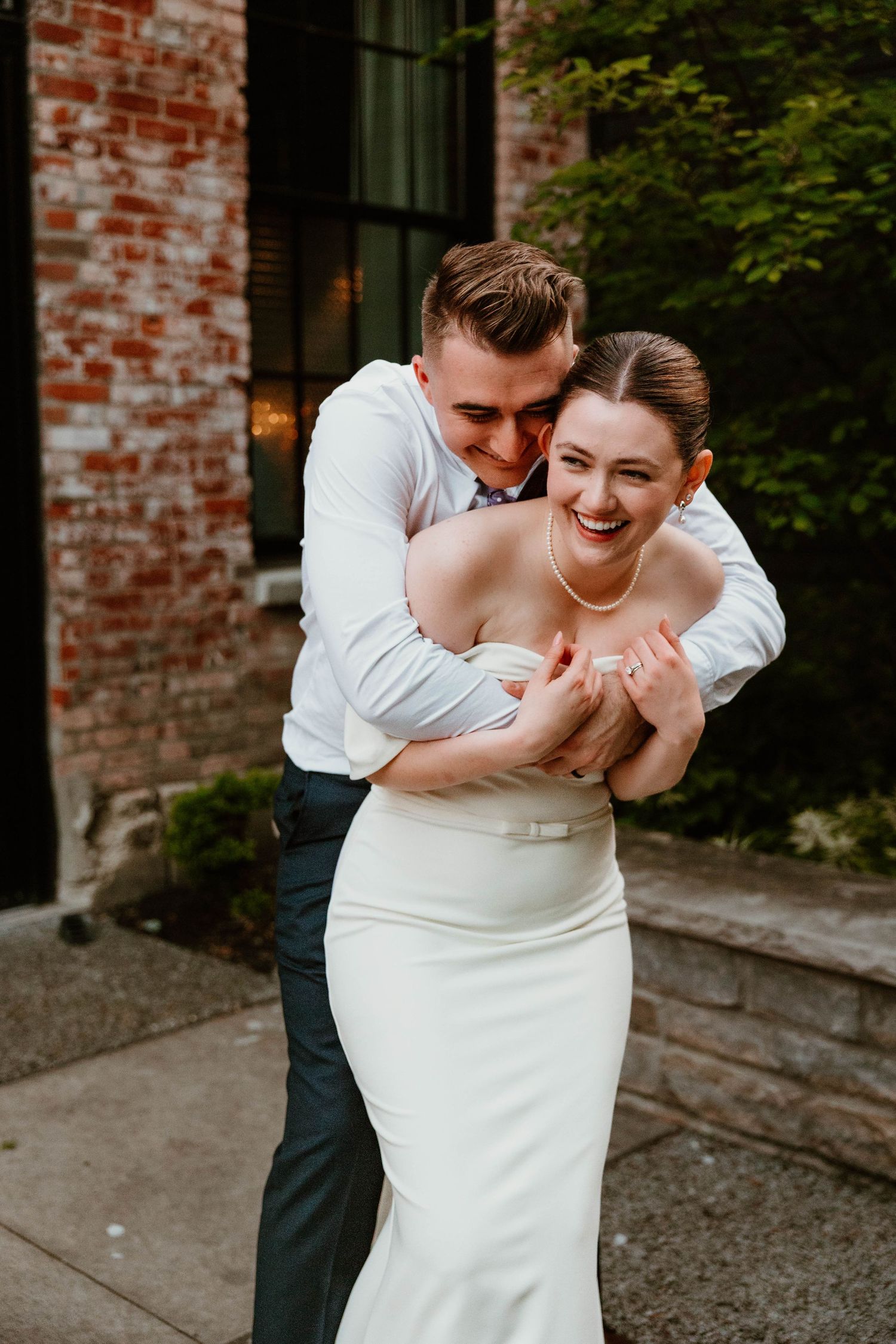 Joyful wedding couple sharing playful embrace outside brick building.