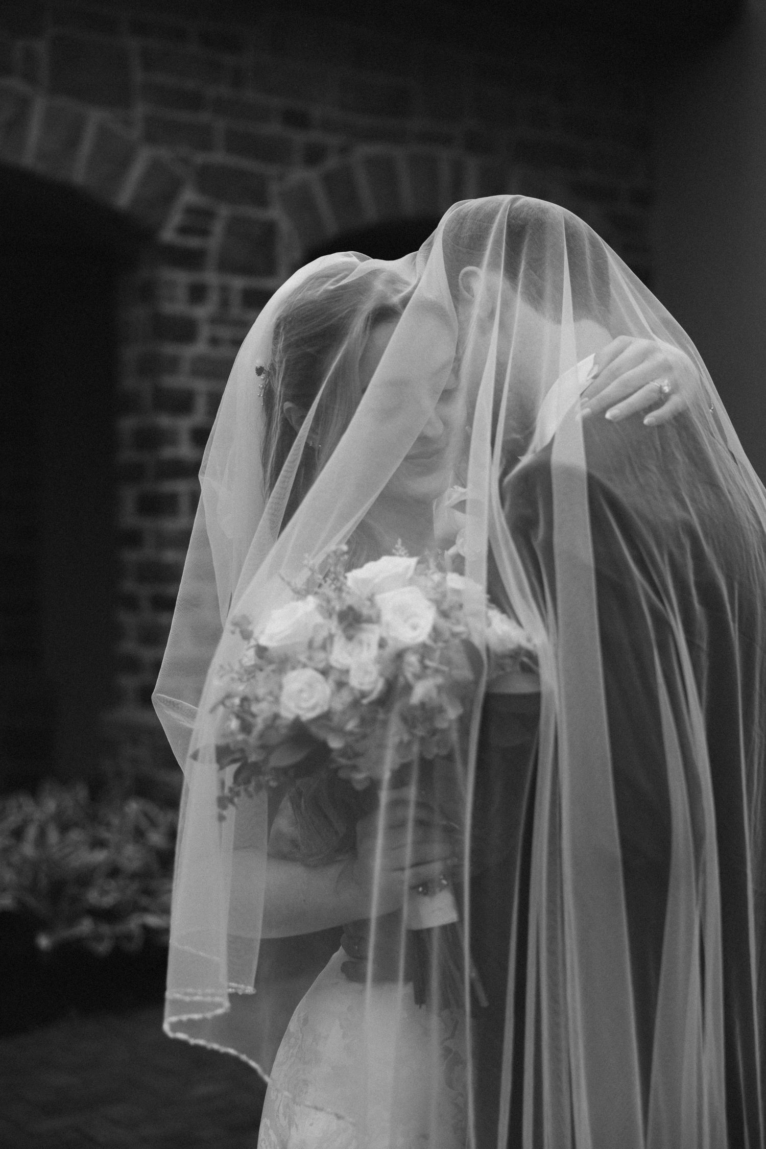 Bride and groom sharing a romantic kiss under a flowing wedding veil, holding a bouquet, in black and white.