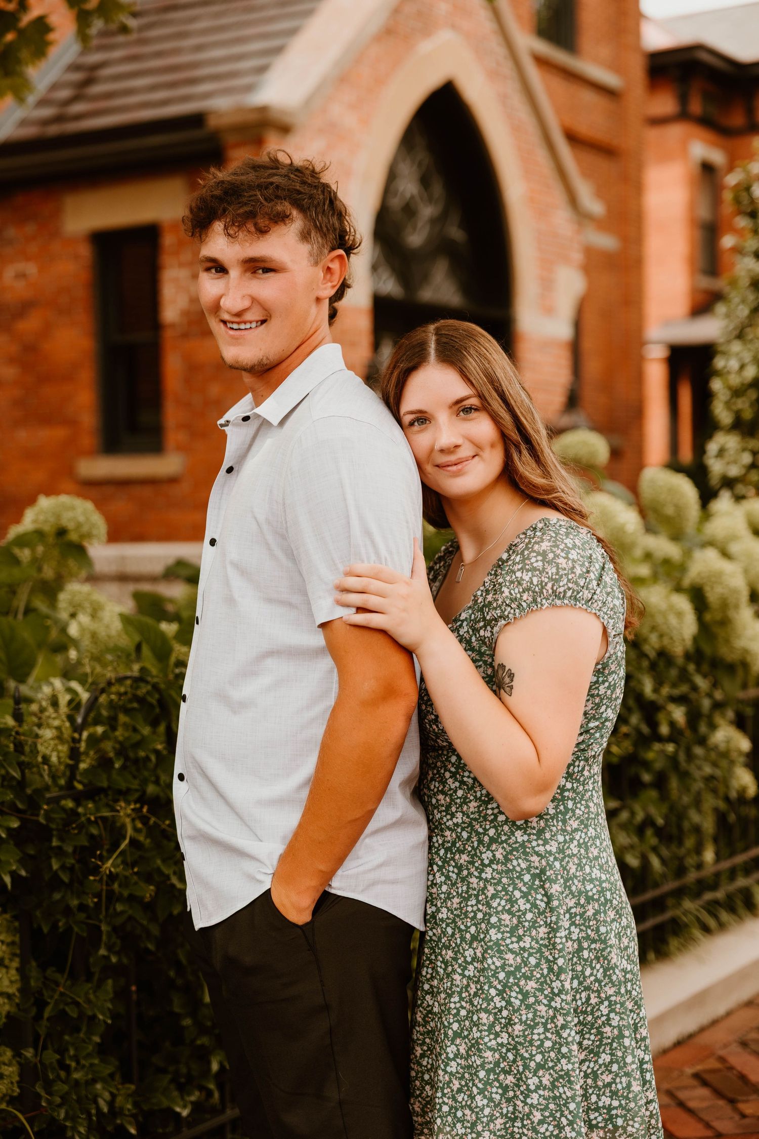 Couple in white shirt and green floral dress share playful moments in front of brick building with garden backdrop.