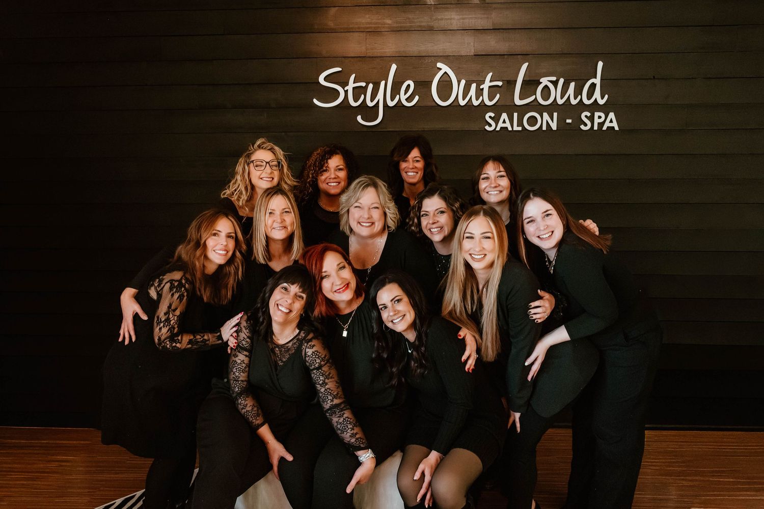 Group photo of salon staff members in black attire against wooden wall with business signage.