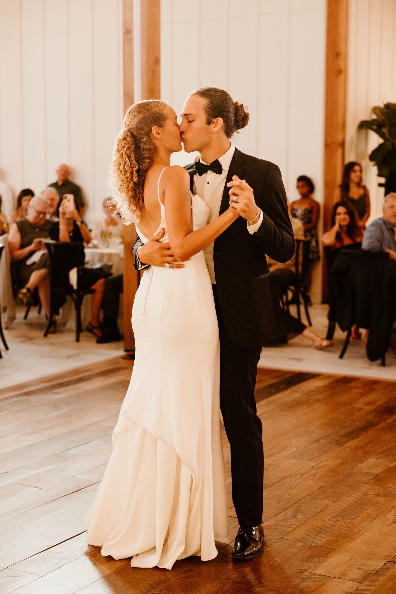 Couples share their first dance in an elegant white-walled reception venue with wooden floors.