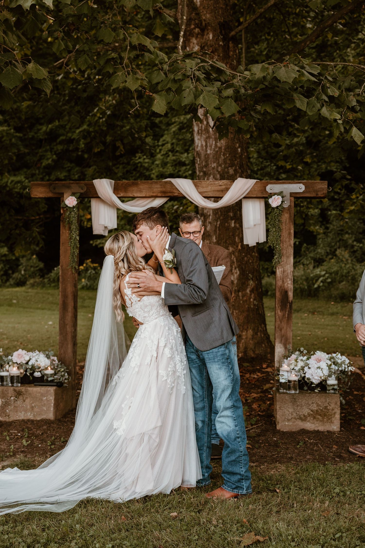 Wedding couple embraces under wooden pergola in outdoor garden setting.