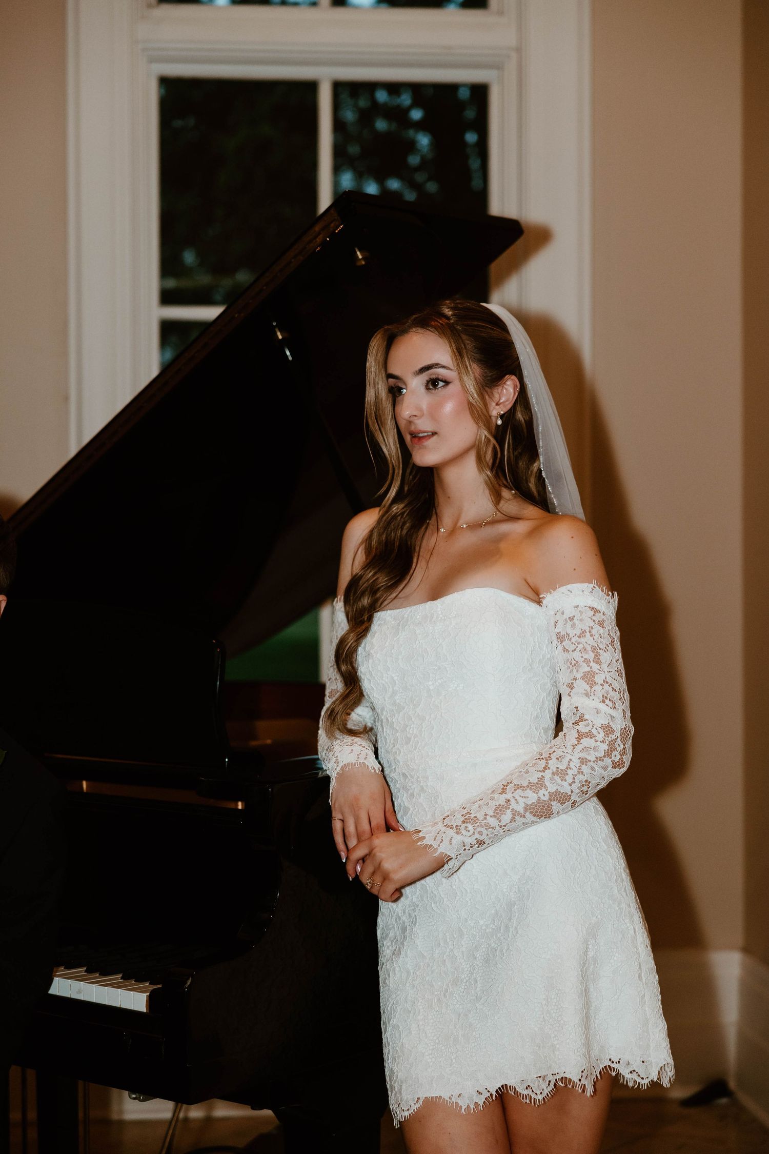 Bride in short white lace off-shoulder dress and veil poses elegantly beside a black grand piano.