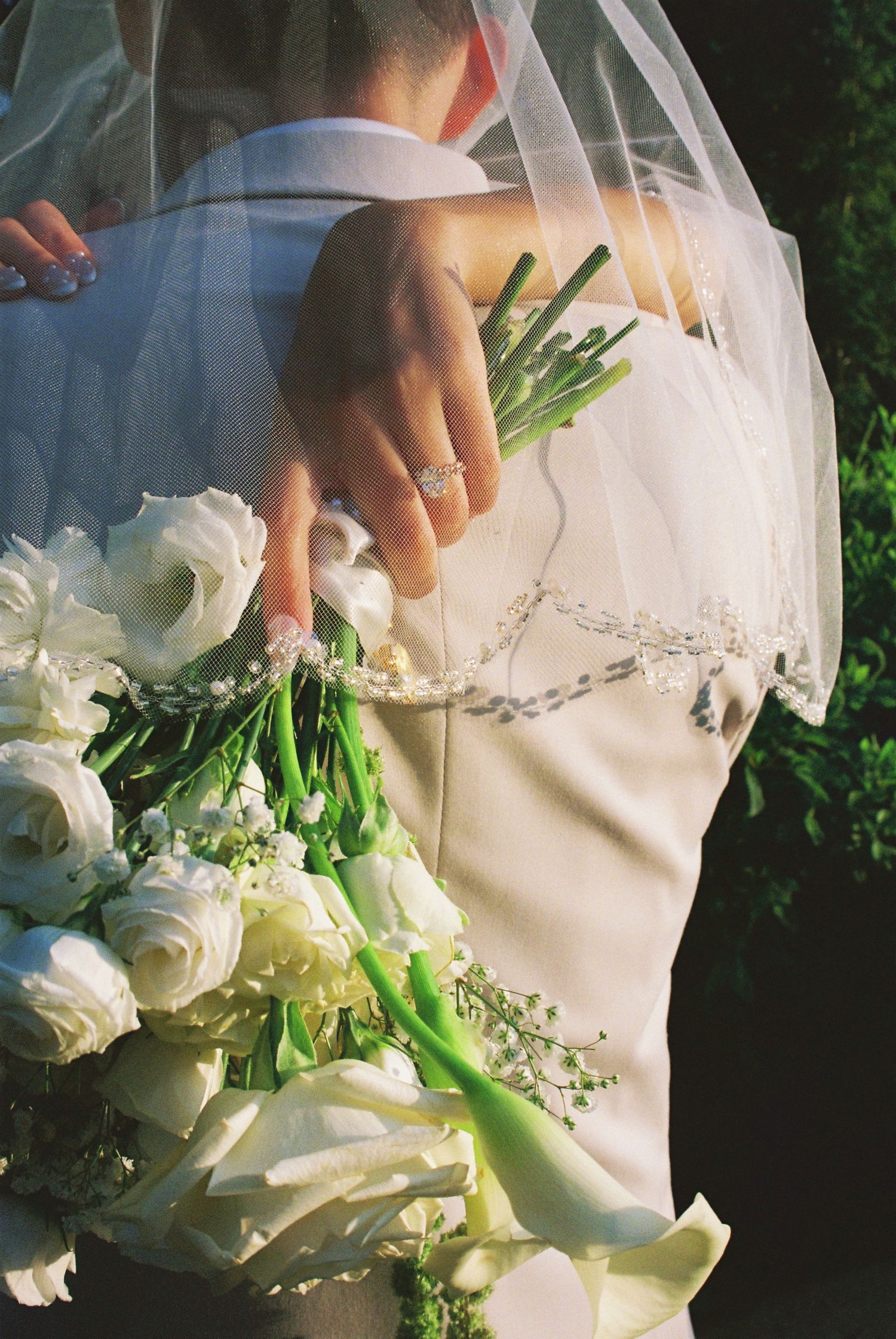 Close-up of bride holding white rose bouquet with groom, showing diamond engagement ring and veil.