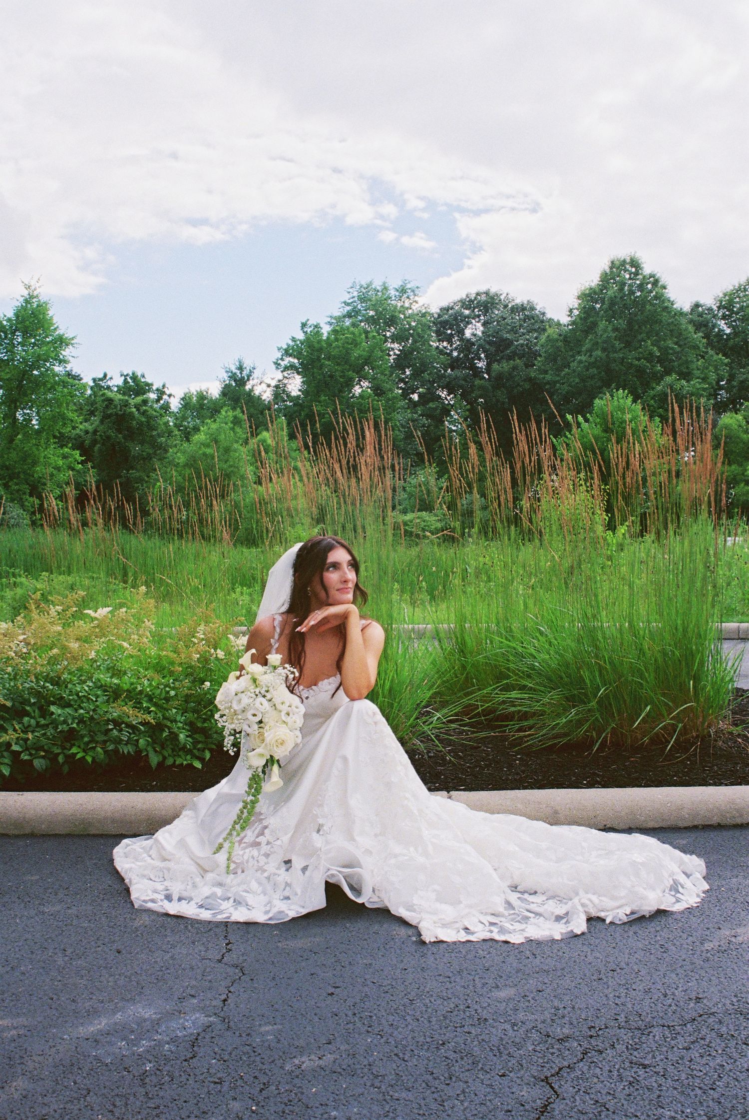 Bride in a lace gown with long train sitting on a curb surrounded by tall ornamental grasses.