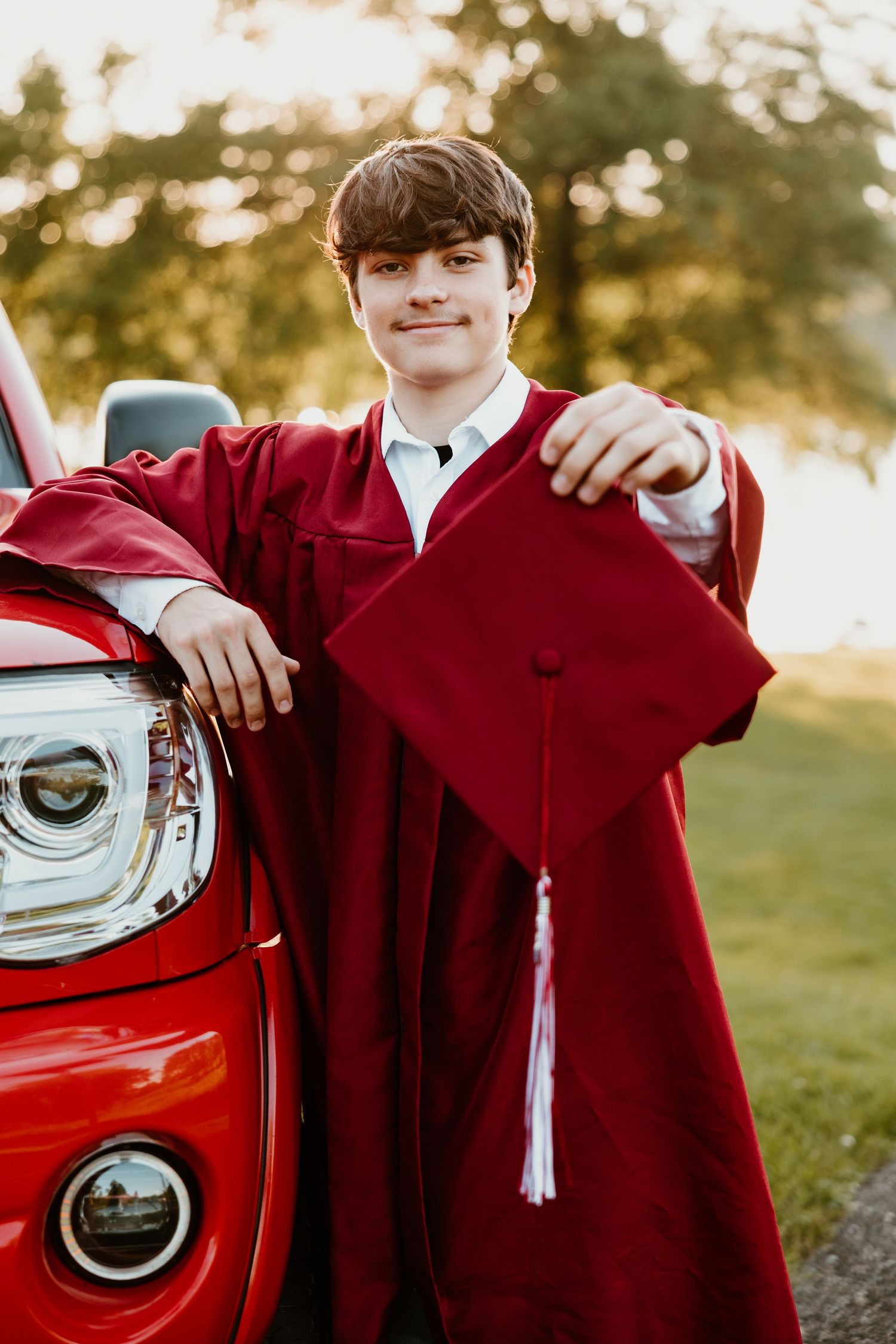Graduate in red cap and gown poses next to red car during sunset.