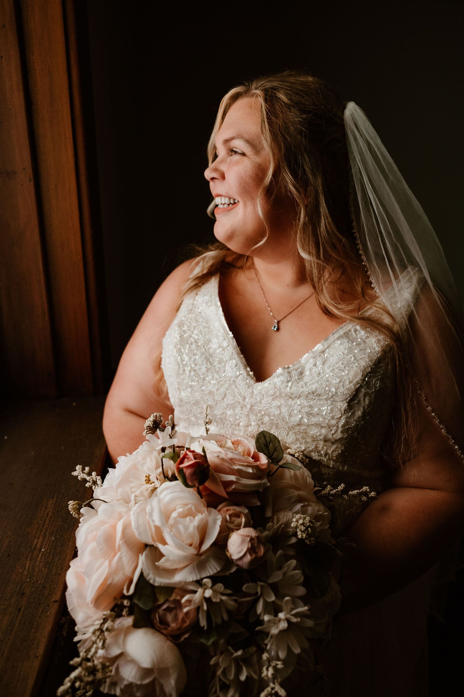 A bride in a white lace dress holds a romantic bouquet of blush and mauve roses against a dark background.