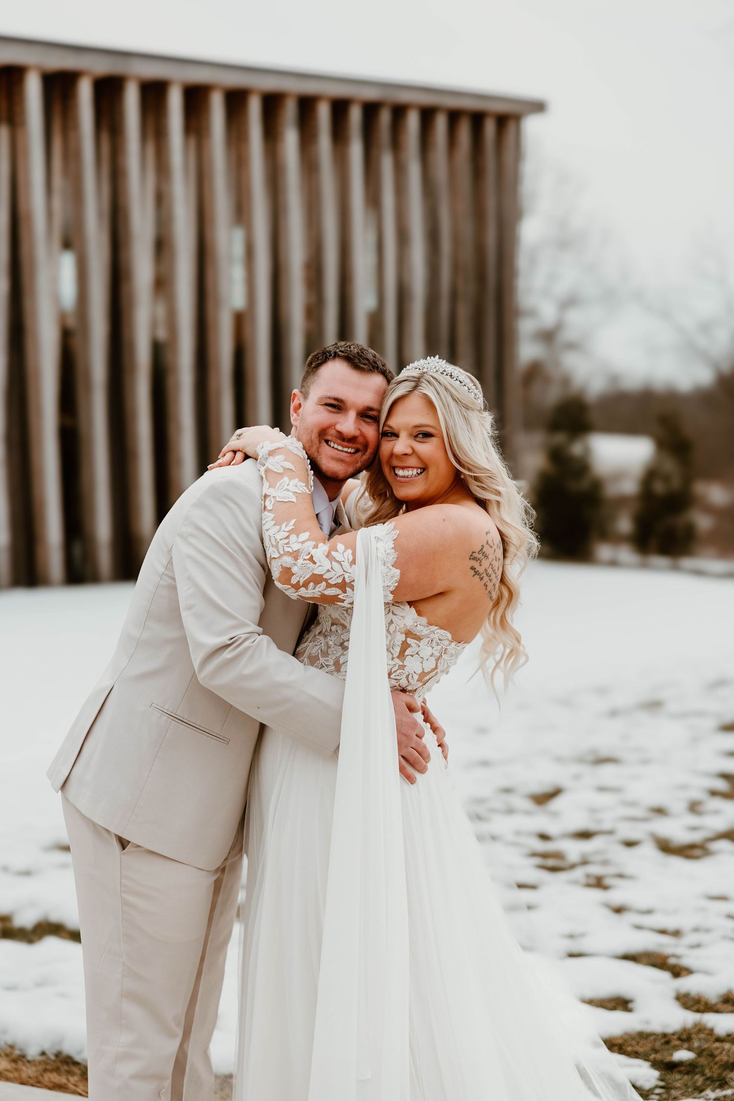A smiling bride and groom embrace in the snow outside a rustic barn, she in a white lace gown and he in a tan suit.