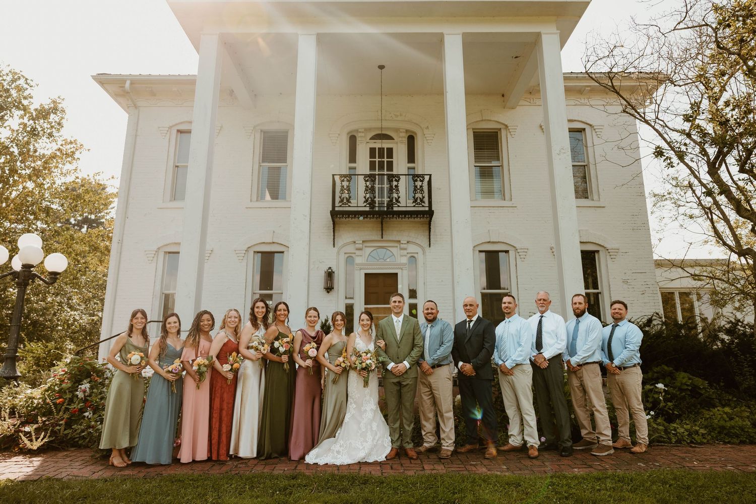 Wedding party poses in front of elegant white Southern mansion with columns, bride and groom centered among bridesmaids and groomsmen.