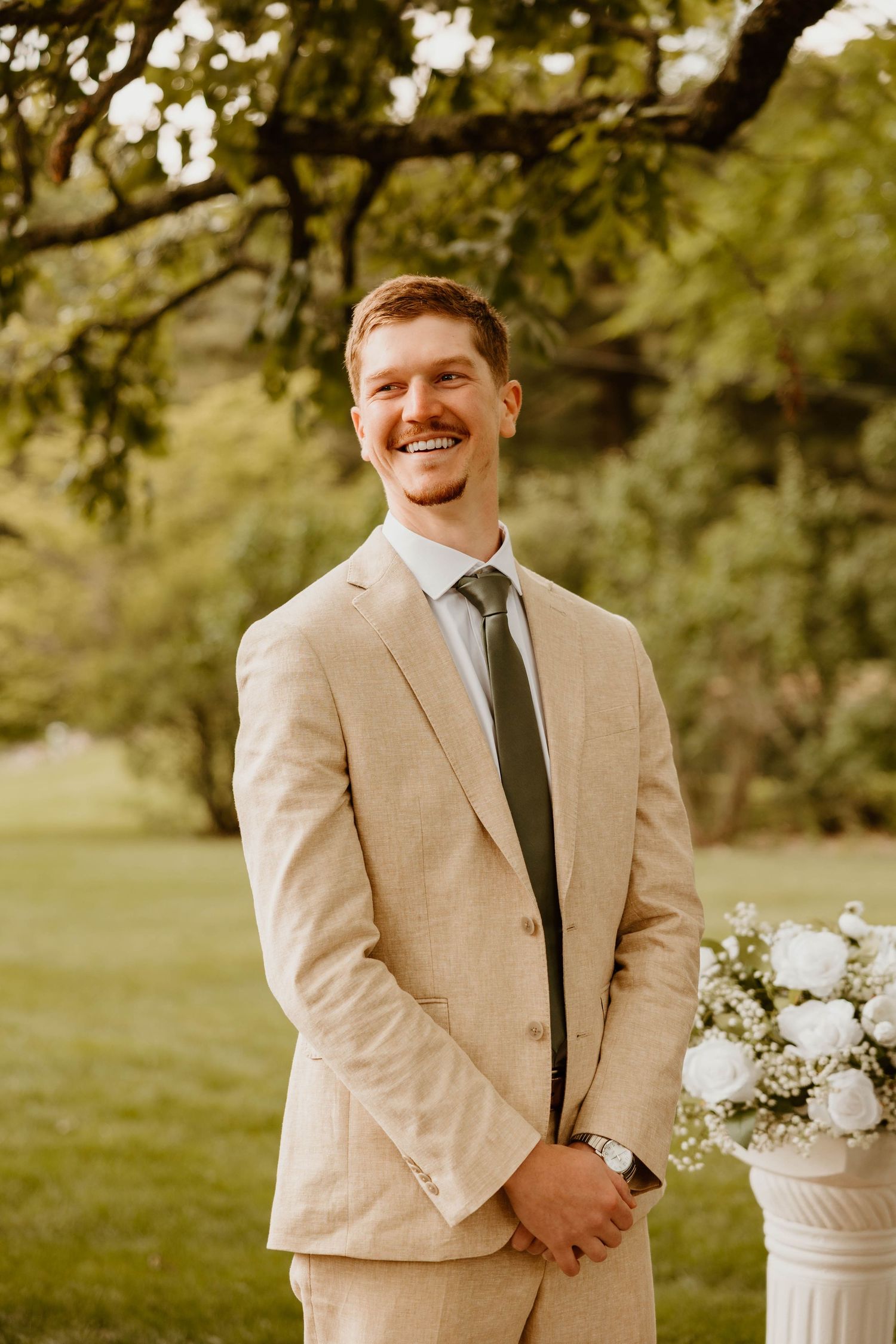 Groom in beige suit and dark tie smiles while standing outdoors during a wedding photoshoot.