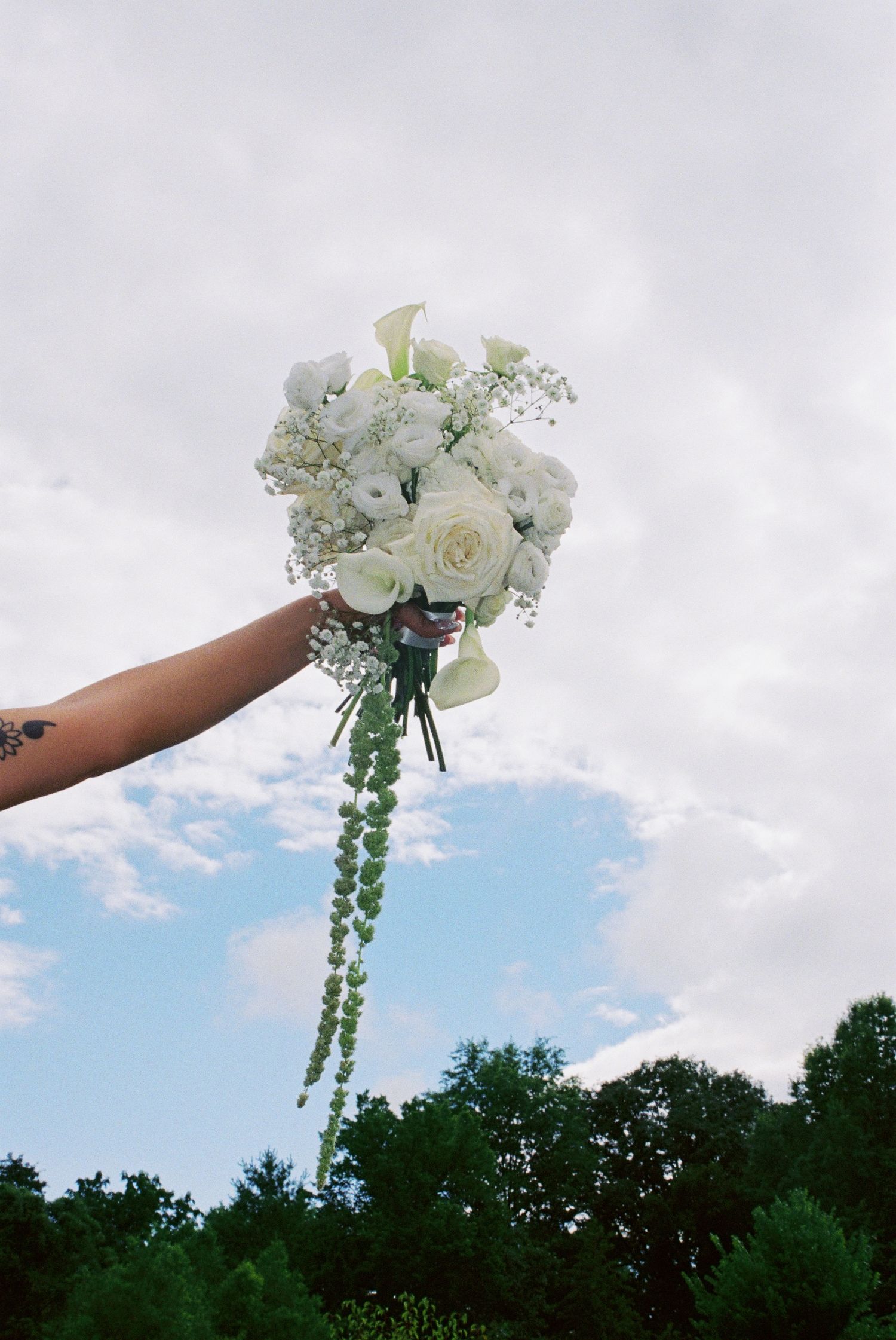 Tattooed arm holding a cascading white rose and calla lily bridal bouquet against a cloudy blue sky.