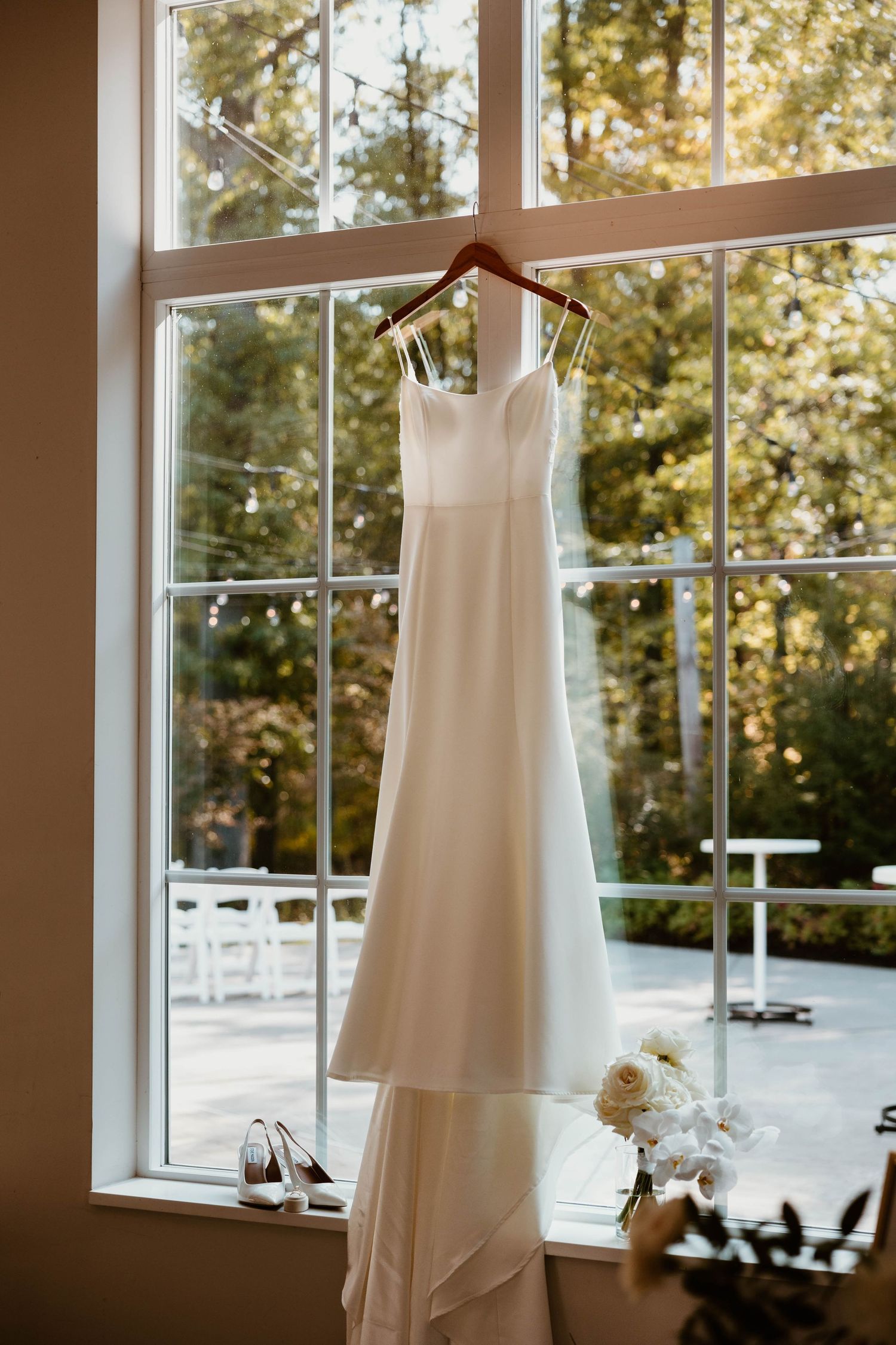Elegant white wedding dress hanging in front of sunlit windows in a bridal suite.