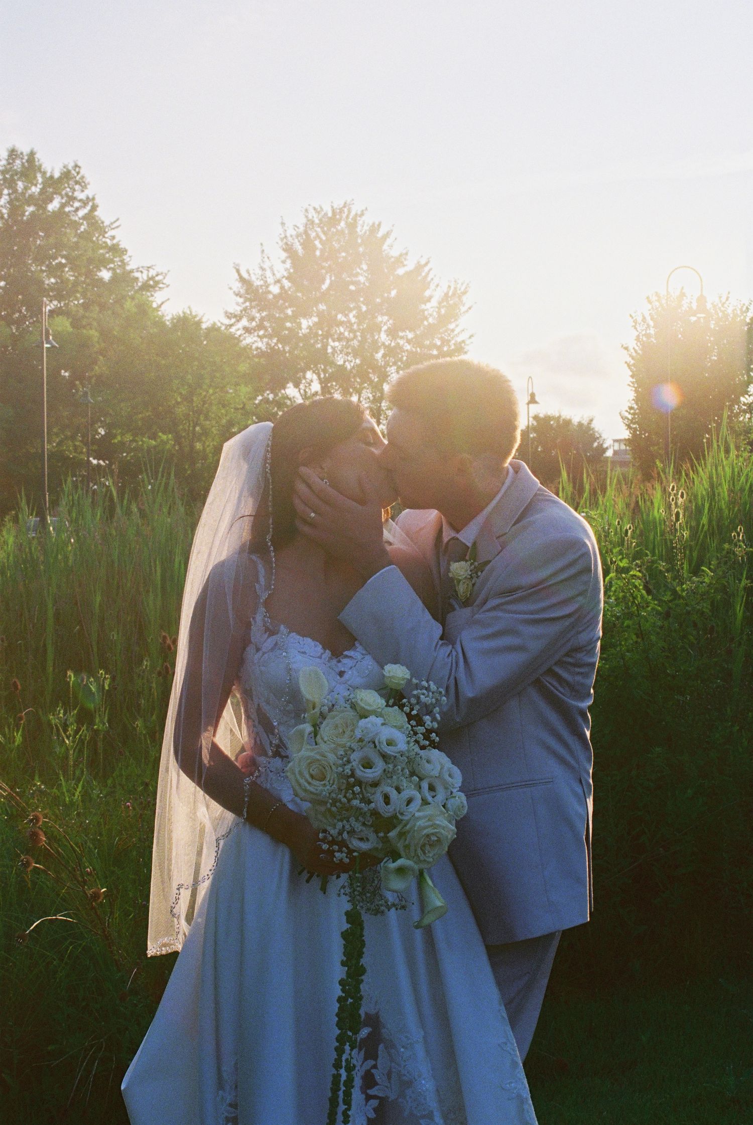 Bride and groom sharing a romantic kiss at sunset in a garden, bride holding white bouquet.