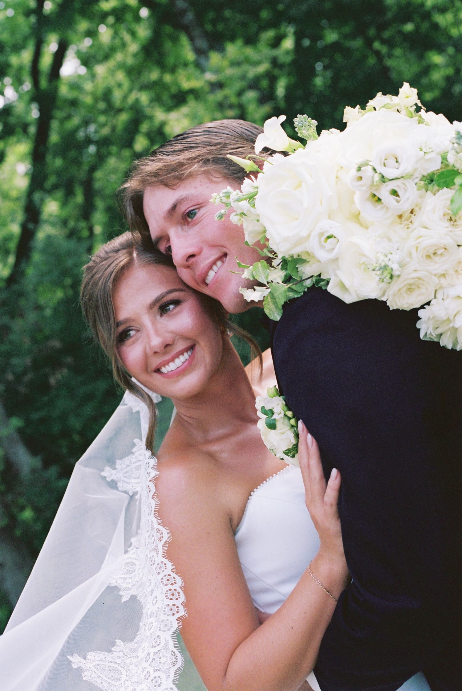 Smiling bride in white gown and veil holds bouquet while groom embraces her outdoors among lush green trees.