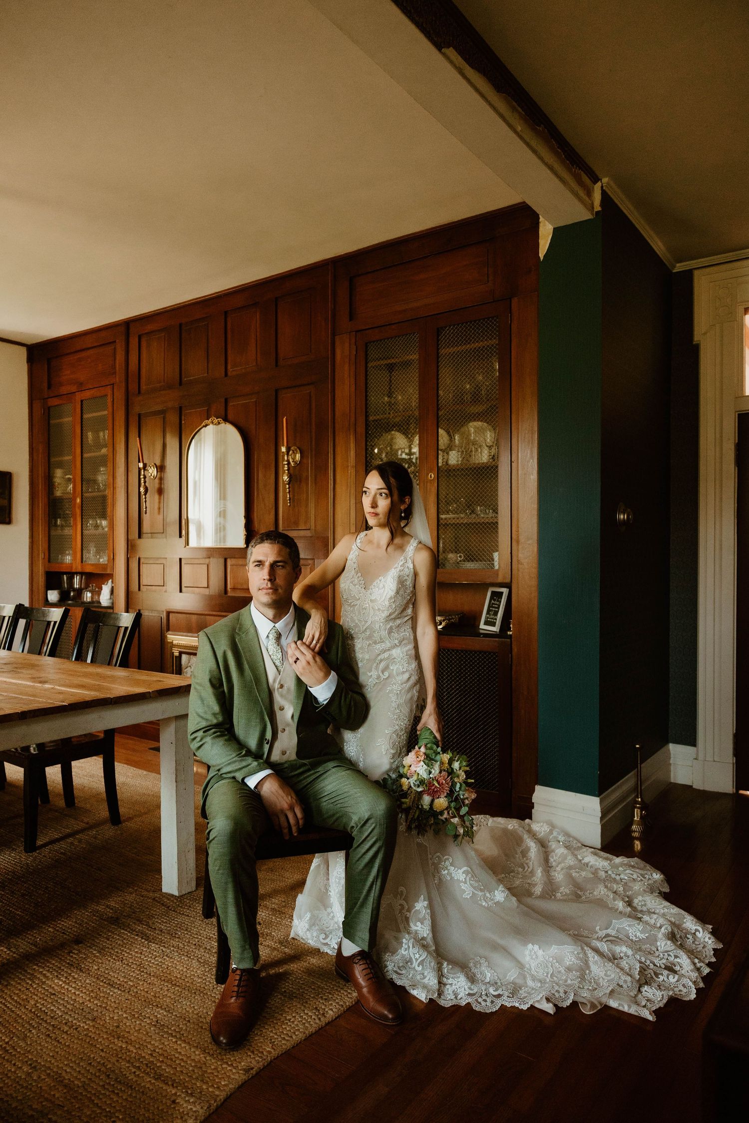 Bride in lace gown holding bouquet stands beside groom in green suit seated in elegant wood-paneled dining room.