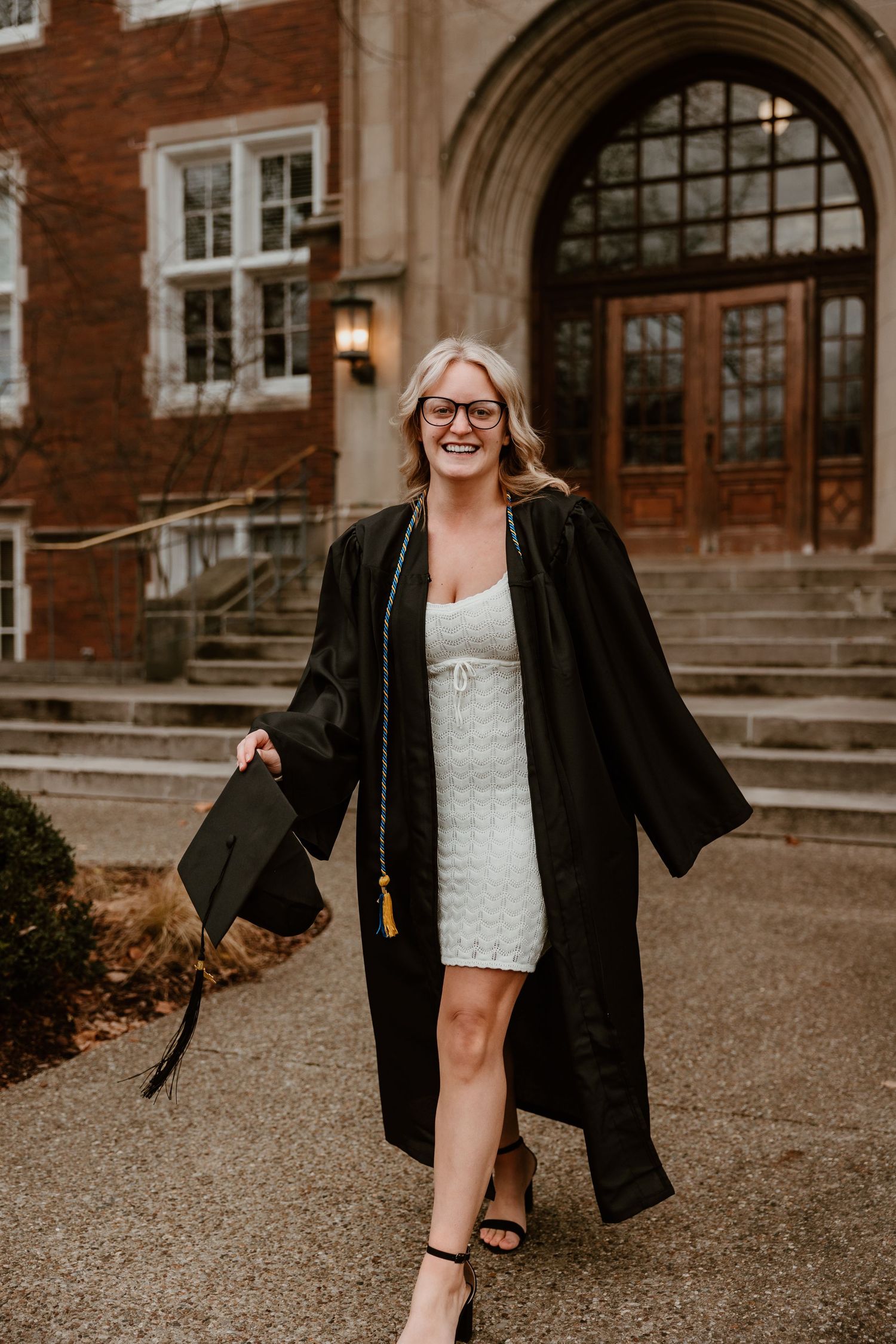 Graduate in black gown poses confidently outside historic brick building entrance.