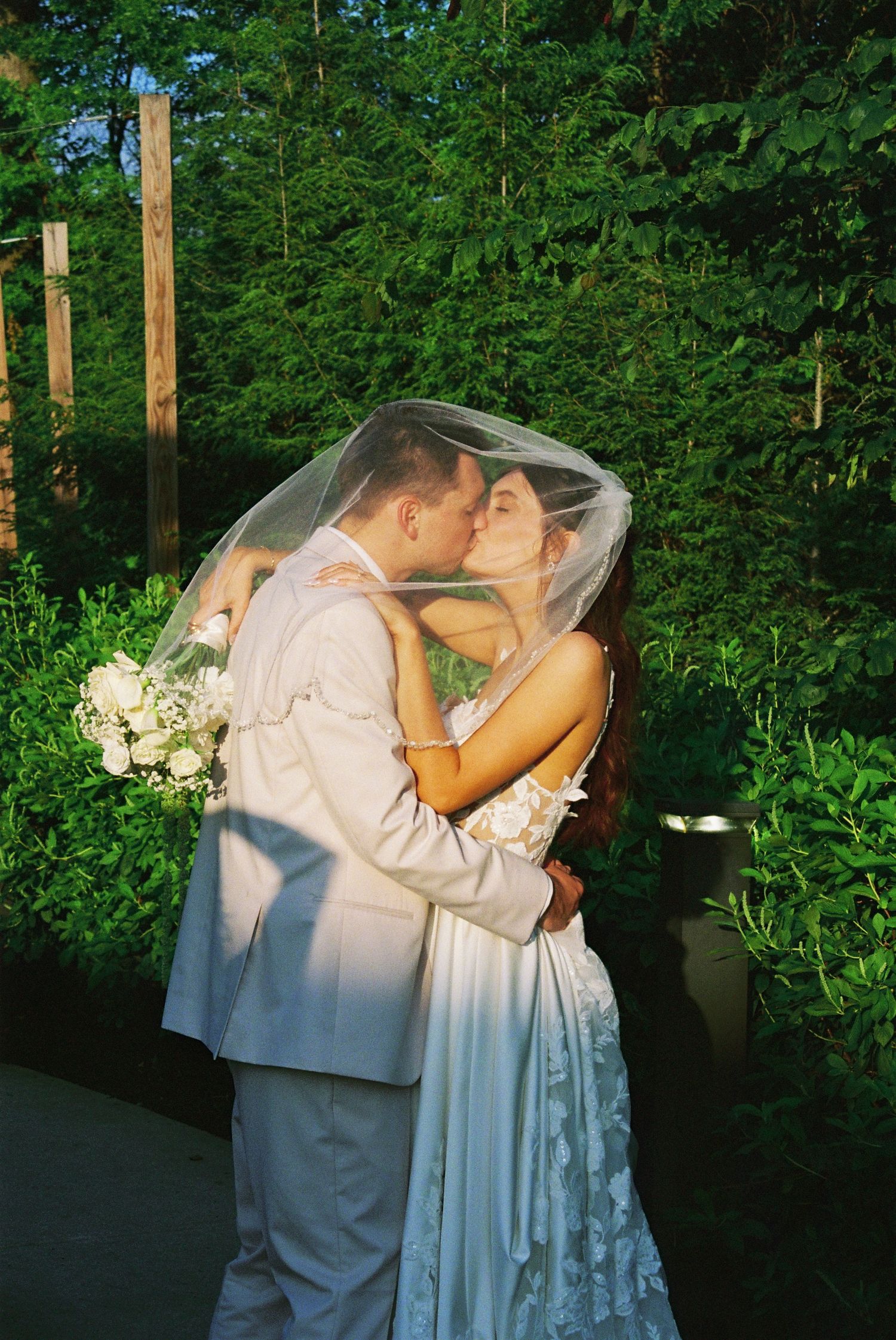 Bride and groom sharing a romantic kiss under a flowing veil in a lush garden setting.