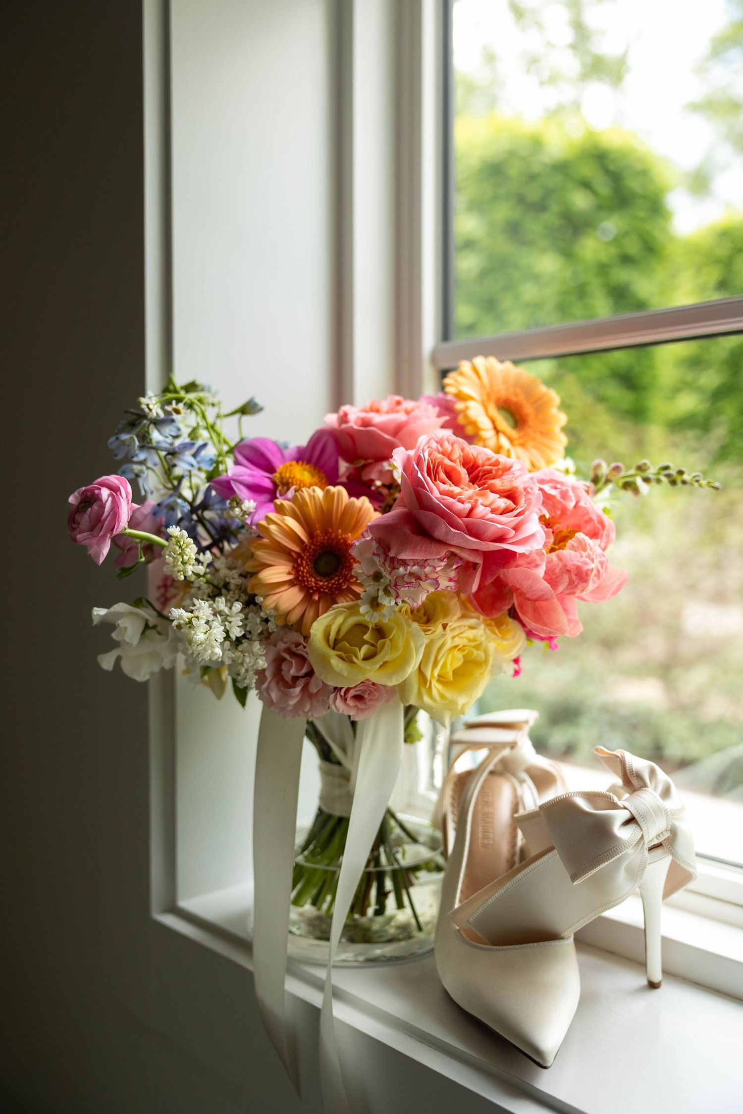 Colorful bridal bouquet with roses and gerberas in a glass vase beside white heels on a sunlit windowsill.