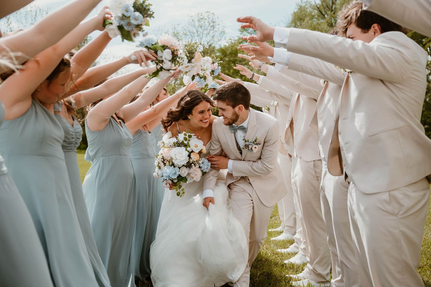 Bride and groom kiss under an arch of raised bouquets held by bridesmaids in blue and groomsmen in white suits.