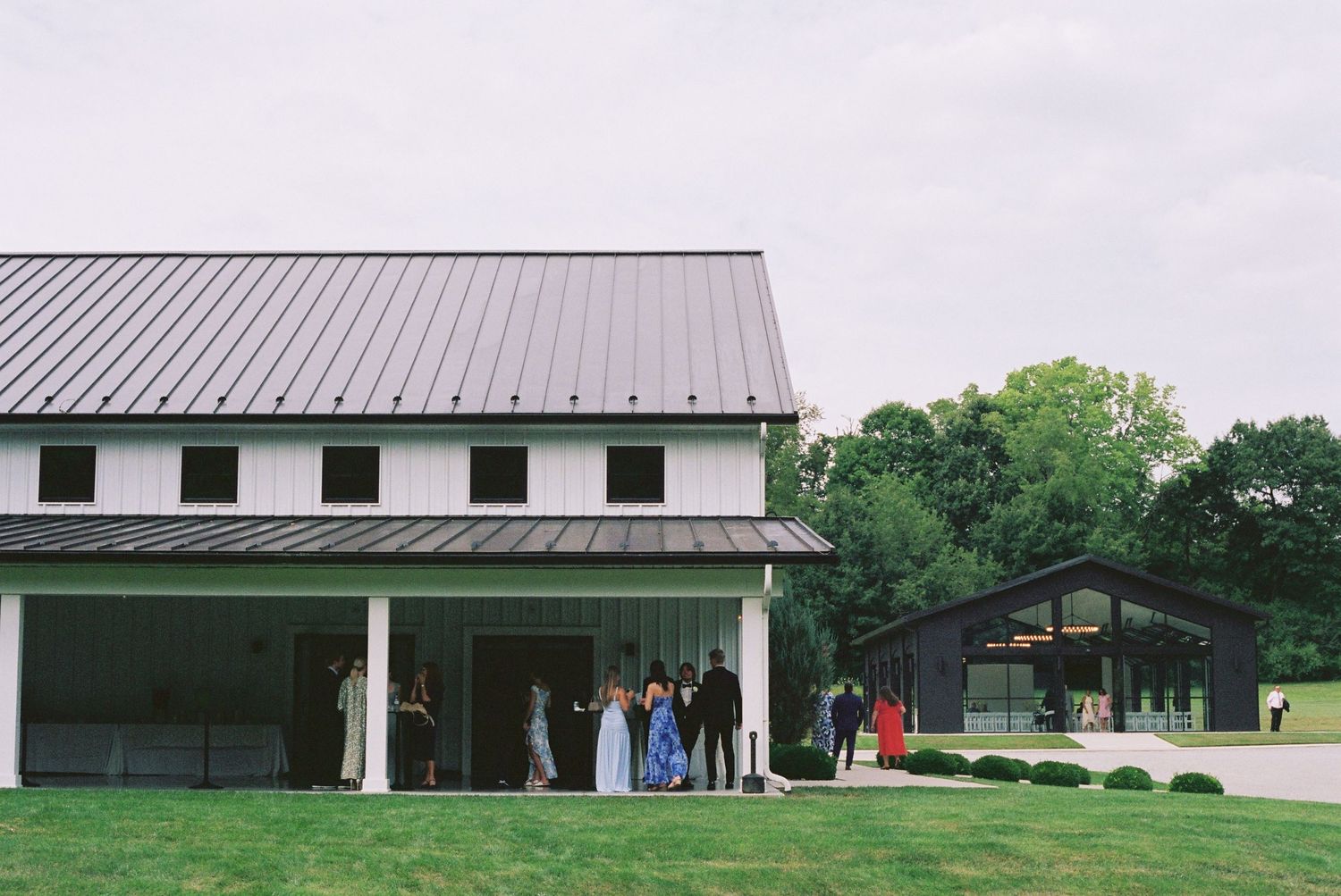 Elegant white barn wedding venue with metal roof, covered porch, and guests gathered on lush green lawn.