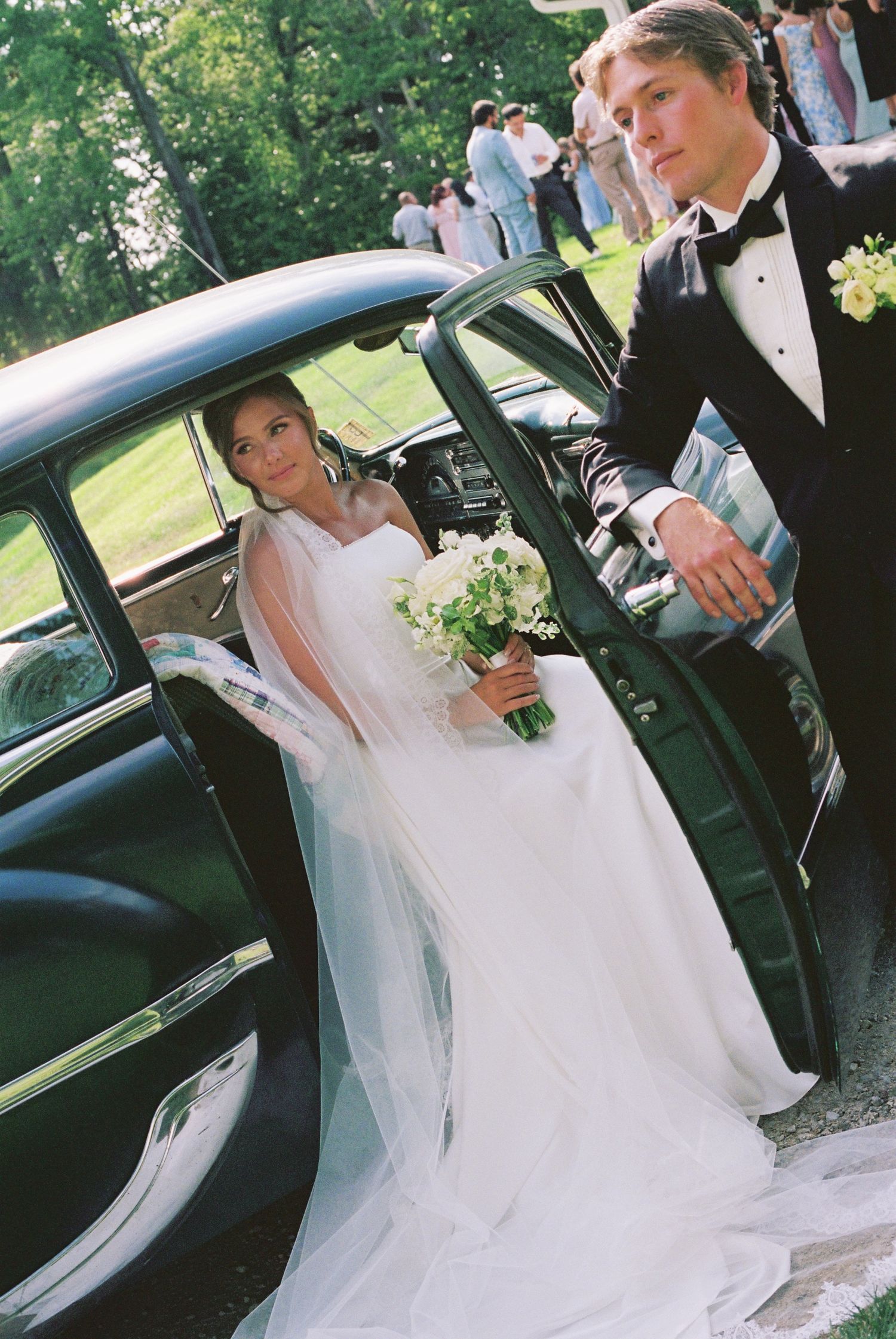 Bride in white gown holding bouquet exits vintage black car as groom in tuxedo holds door open outdoors.