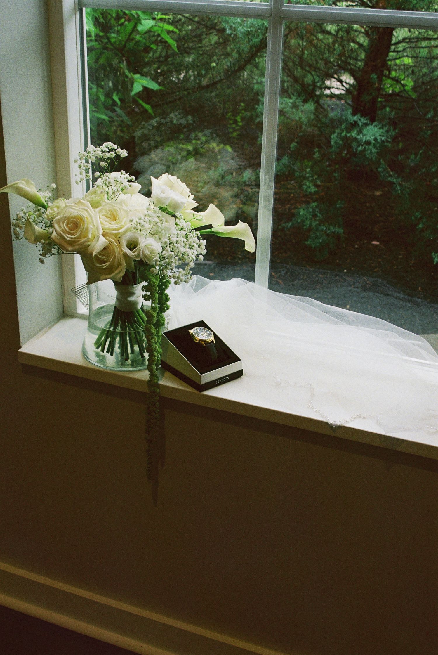 Bridal bouquet of white roses and calla lilies beside a ring box on a windowsill with veil.