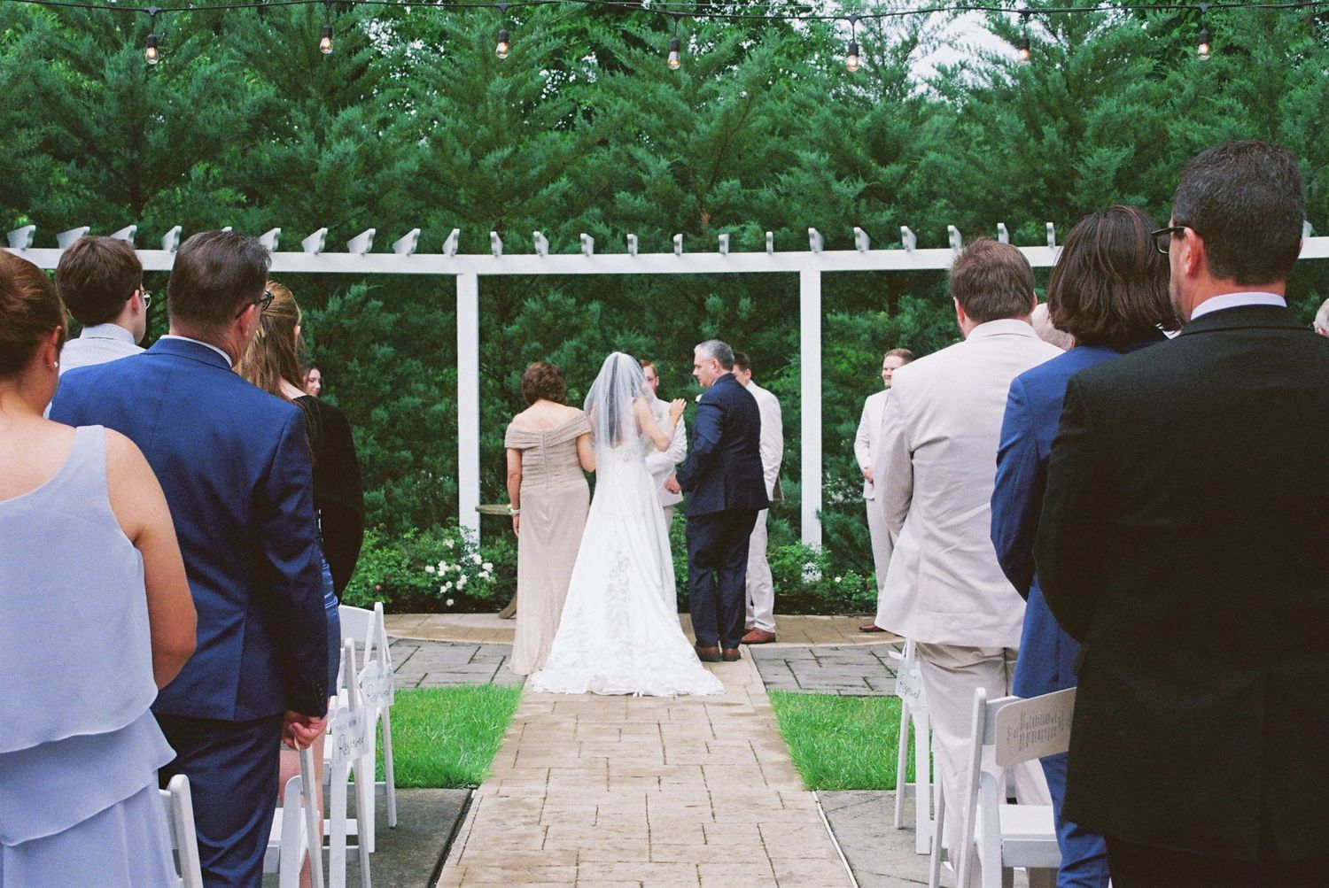 Outdoor wedding ceremony with bride and groom at white pergola altar surrounded by seated guests.