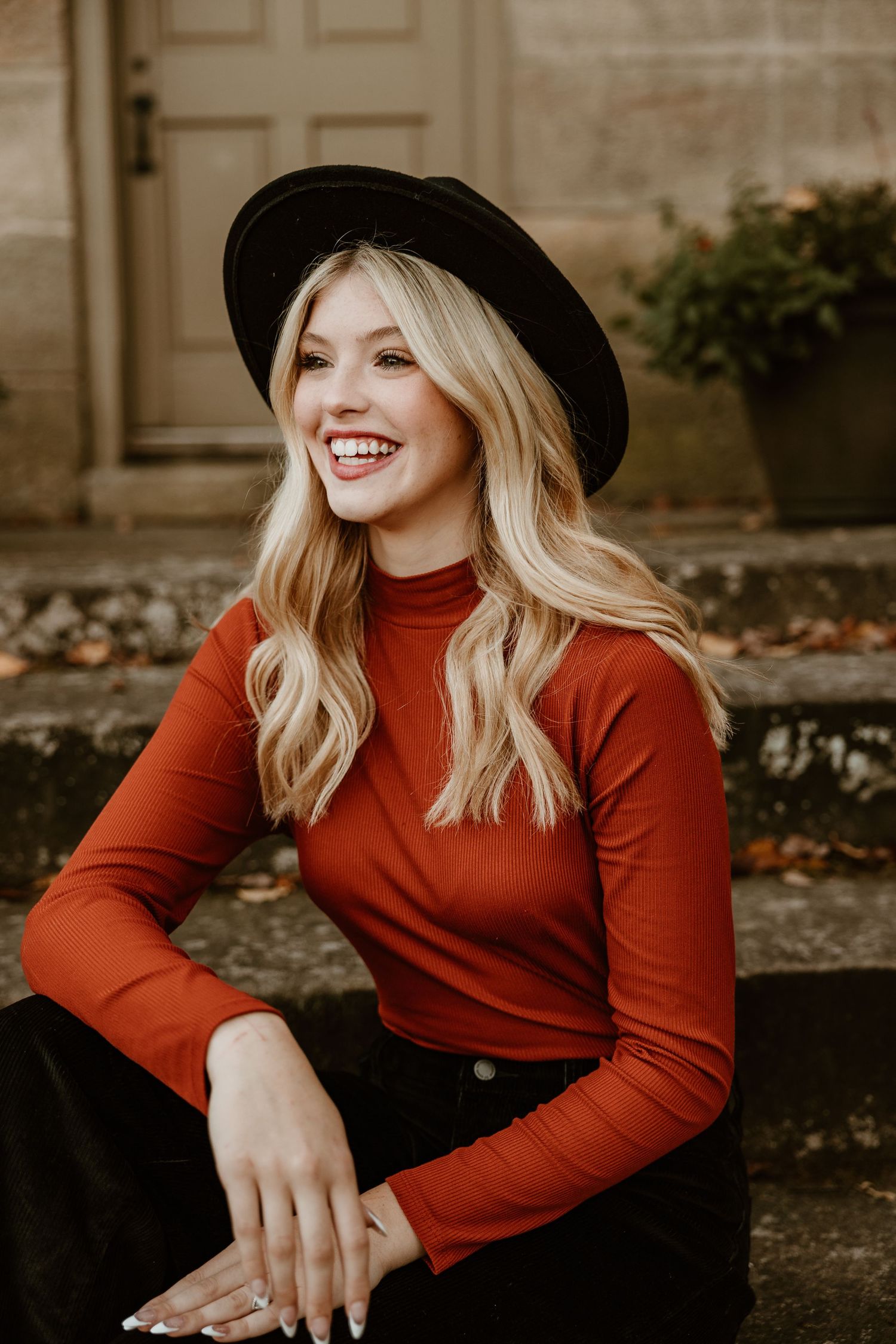 Woman in red turtleneck and black hat poses against stone wall during fall season.