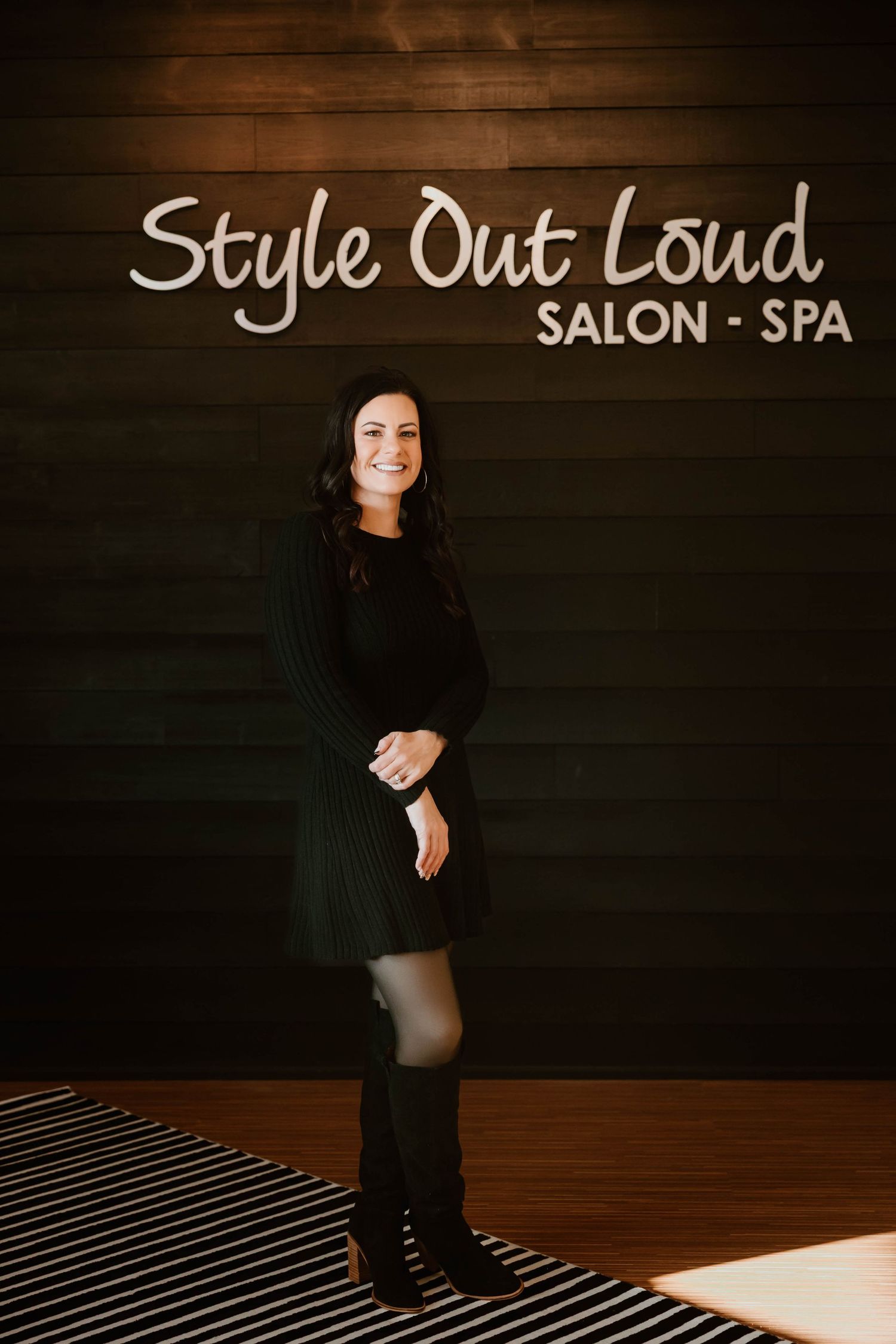 Full-length portrait in black attire against wooden wall with salon branding.