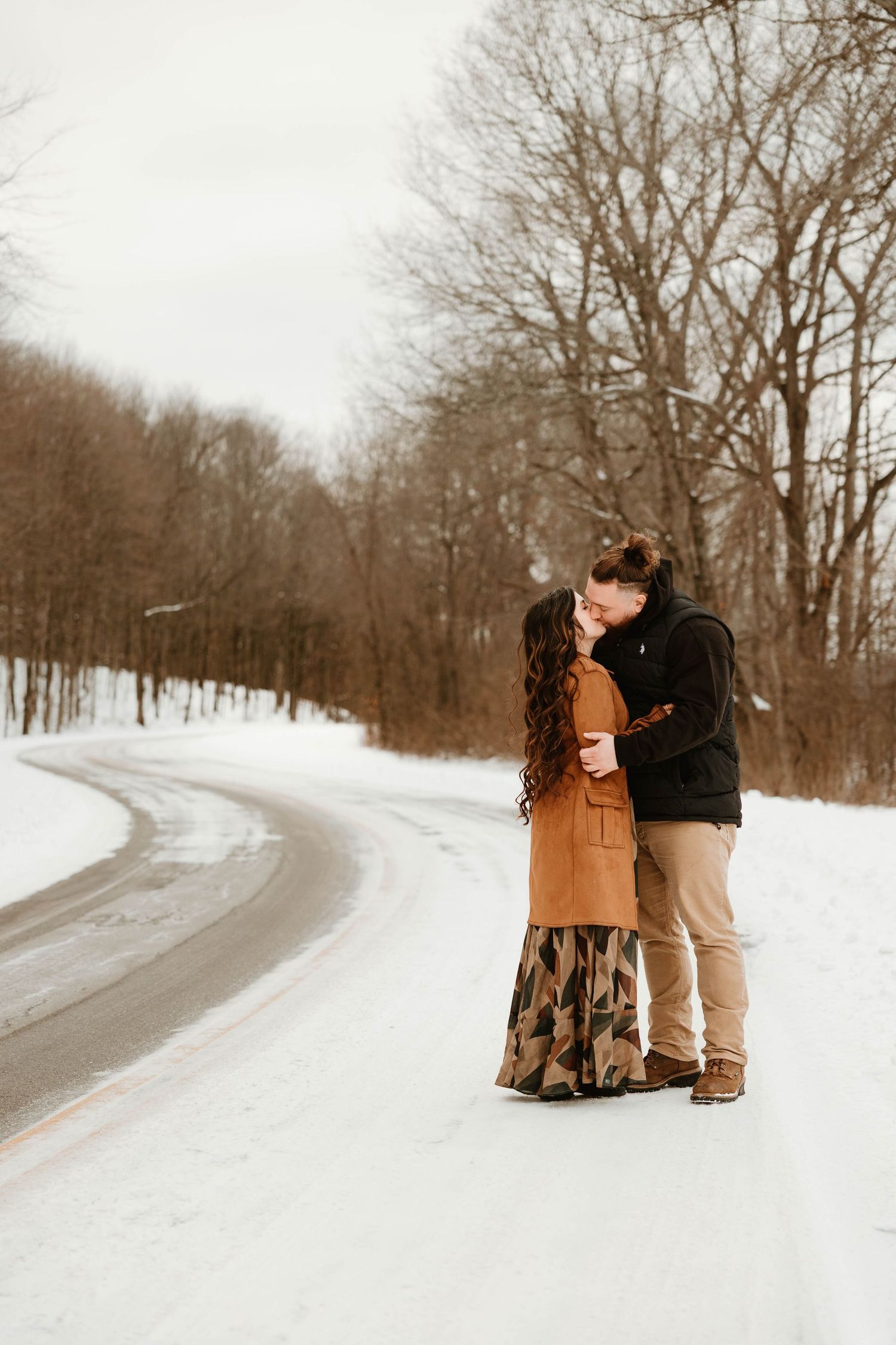 Couple embracing on a snowy winter road surrounded by bare trees.