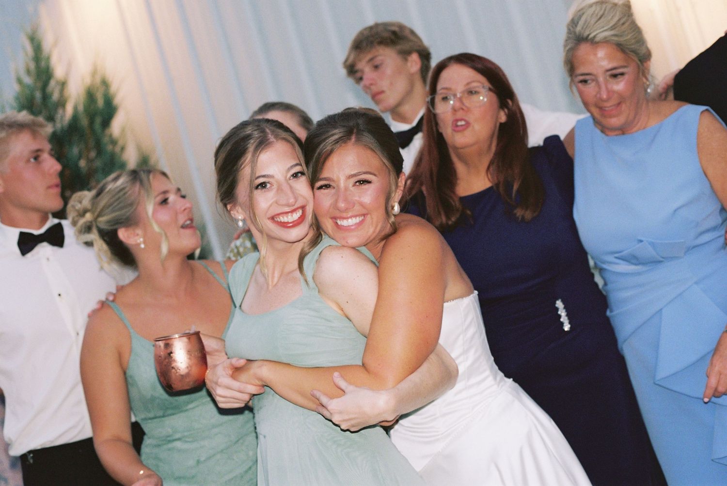 Bride in white gown hugging bridesmaid in mint dress at wedding reception with guests in background.