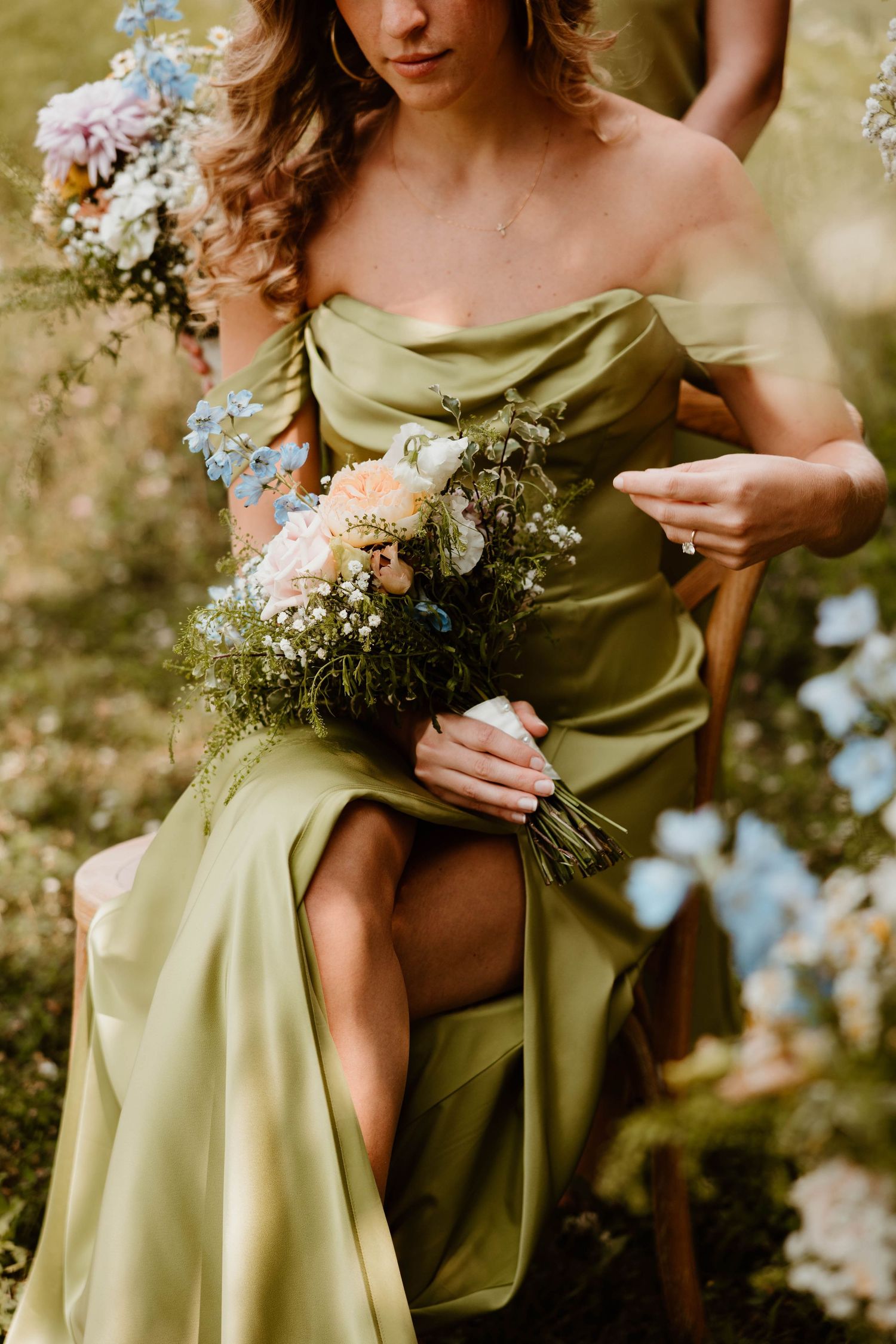 Woman in olive green off-shoulder gown holding a pastel wildflower bouquet while seated outdoors.